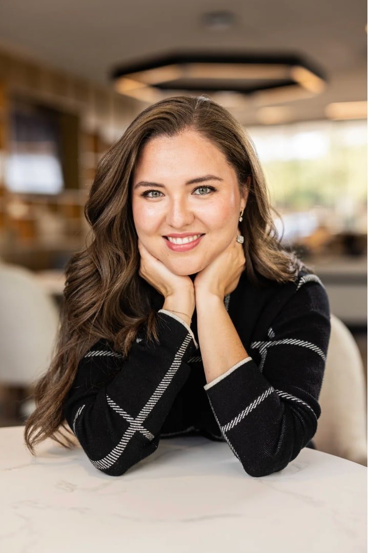 Business portrait of a smiling woman in a black jacket leaning on a chair in a softly blurred interior.