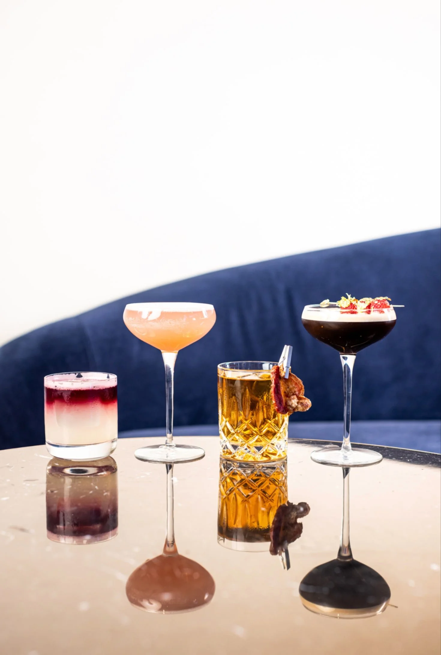 Four colorful cocktails in different glasses on a reflective table beside a blue banquette.