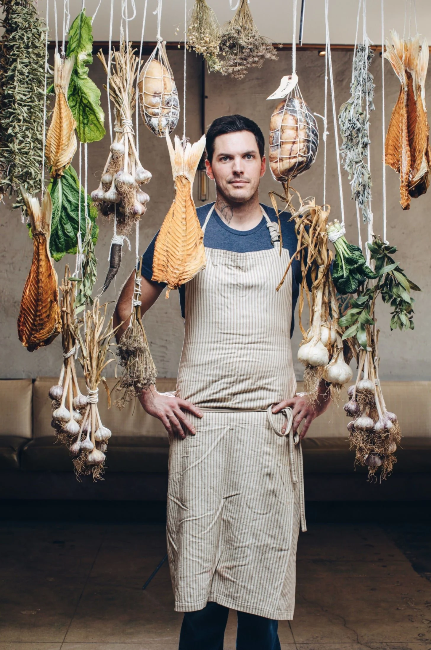 Chef portrait with an apron and hanging bundles of corn, garlic, and greenery.
