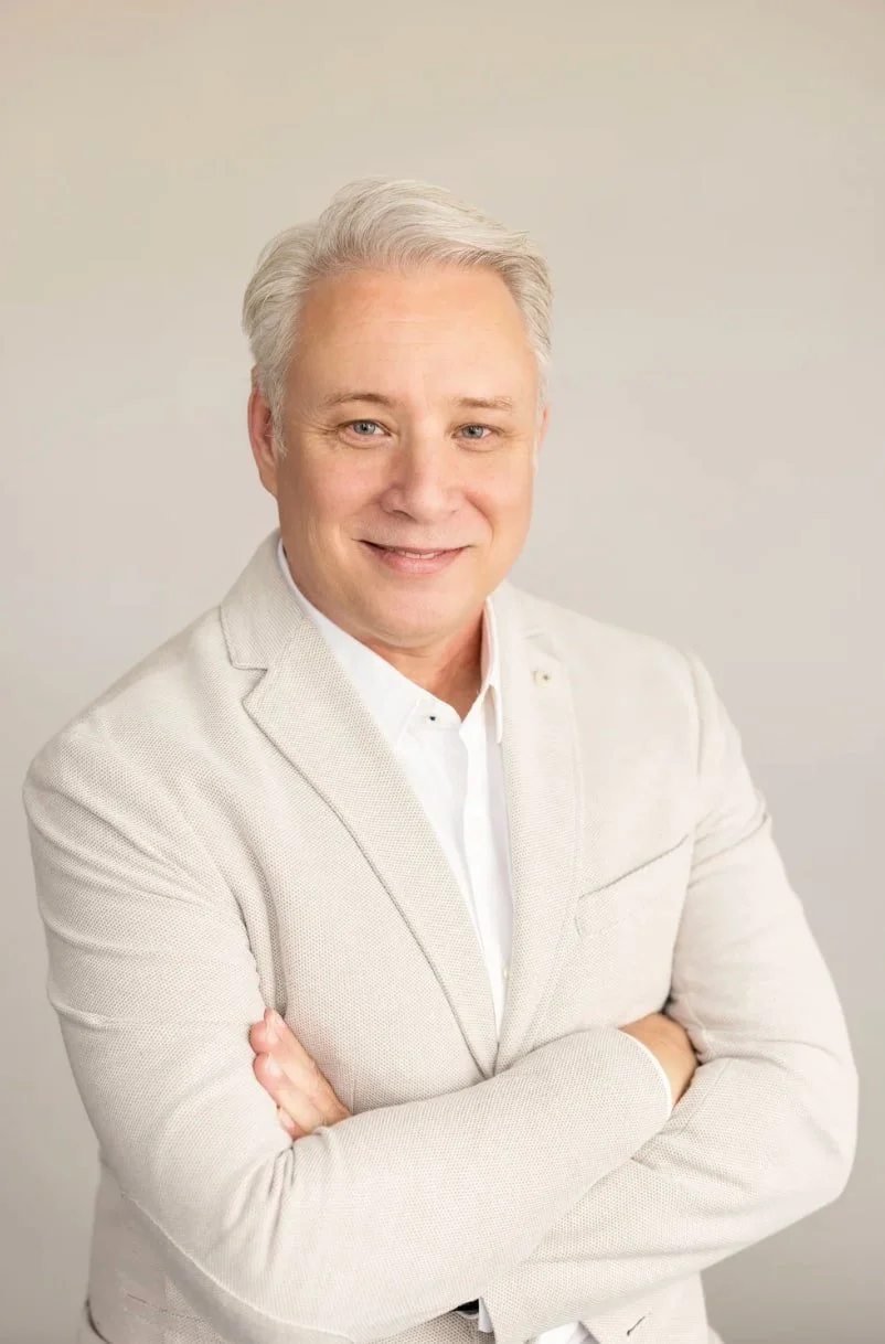 Studio headshot of a smiling man in a light-colored jacket against a neutral beige background.