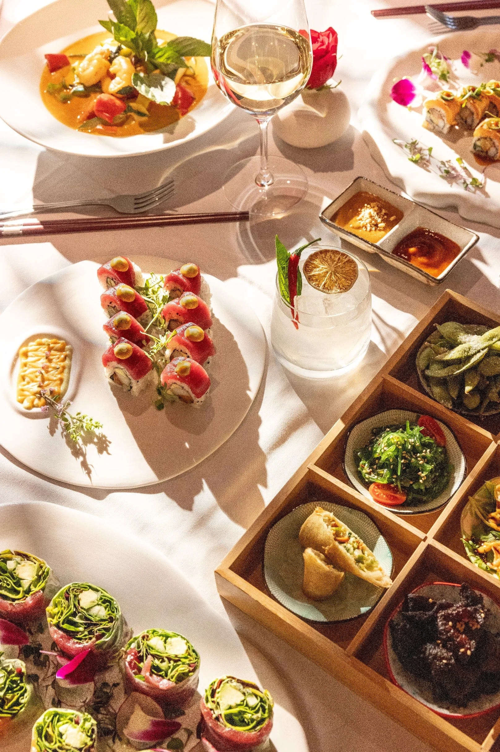 Overhead food photograph of colorful plated dishes, garnishes, and a cocktail on a sunlit restaurant table.