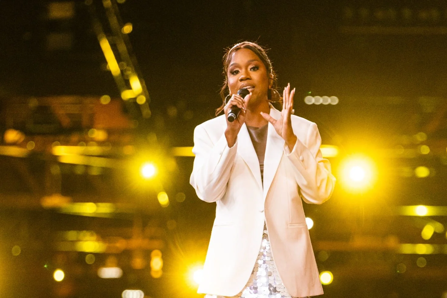 Woman in a white suit speaking into a microphone on a warmly lit event stage.