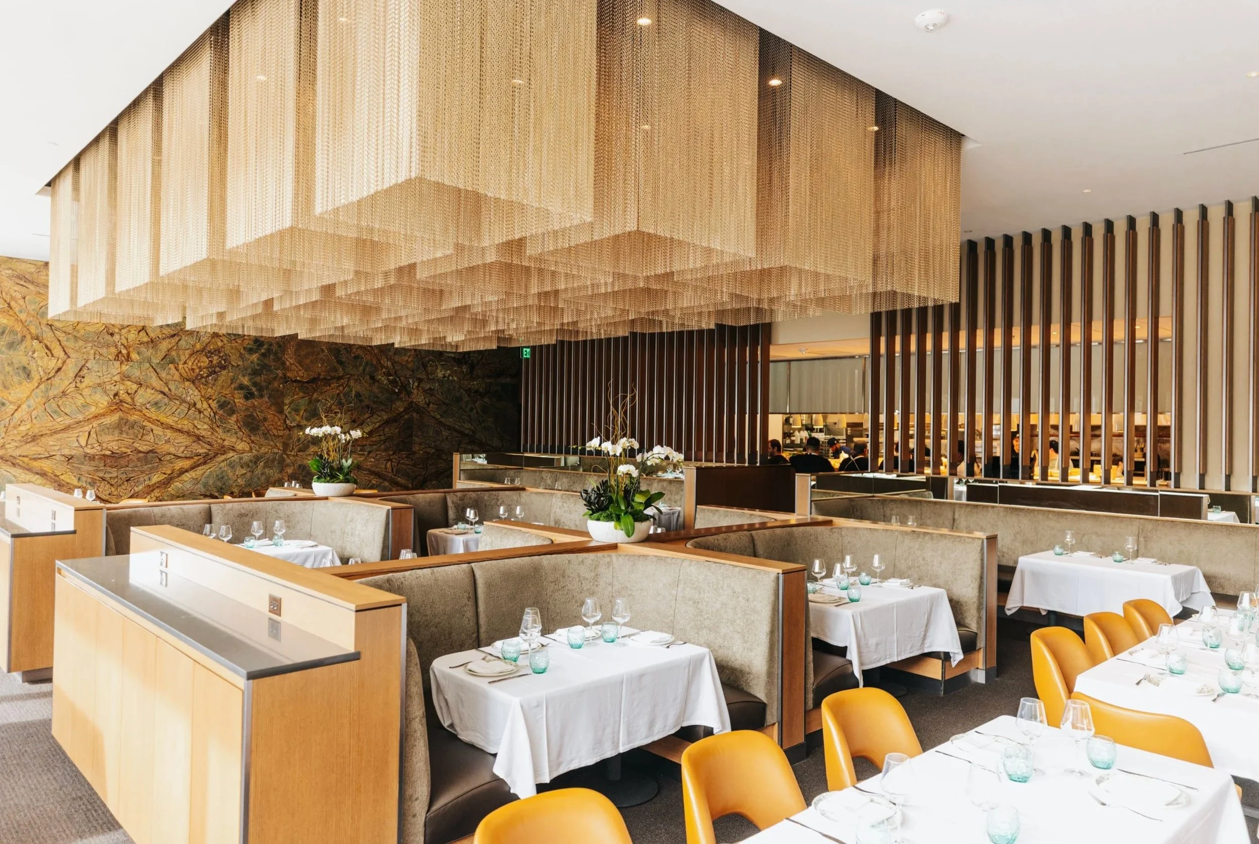 Restaurant dining room with white tablecloths, booth seating, and a sculptural gold-toned ceiling installation.