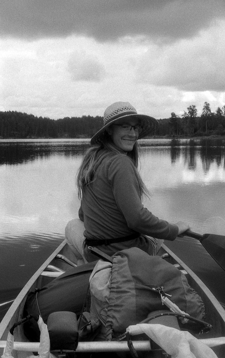 A woman with long hair, wearing glasses, a wide-brimmed hat, and a jacket, is sitting in a small boat on a calm lake, smiling at the camera during daytime with cloudy skies and forested shoreline in the background.