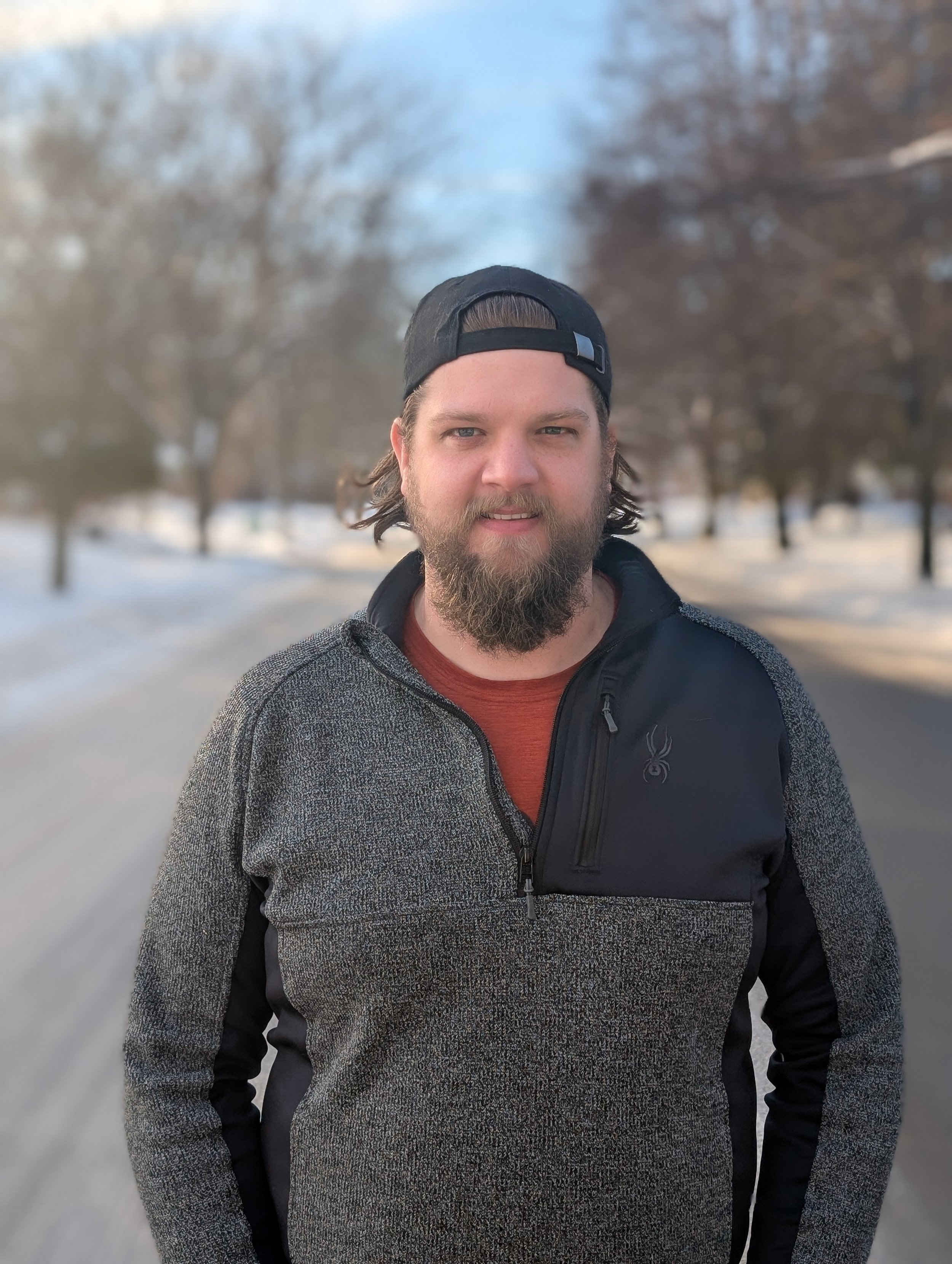 A man with a beard and long hair, wearing a black backward cap, a gray and black jacket, and an orange shirt, standing outdoors on a winter day with snow-covered ground and leafless trees in the background.