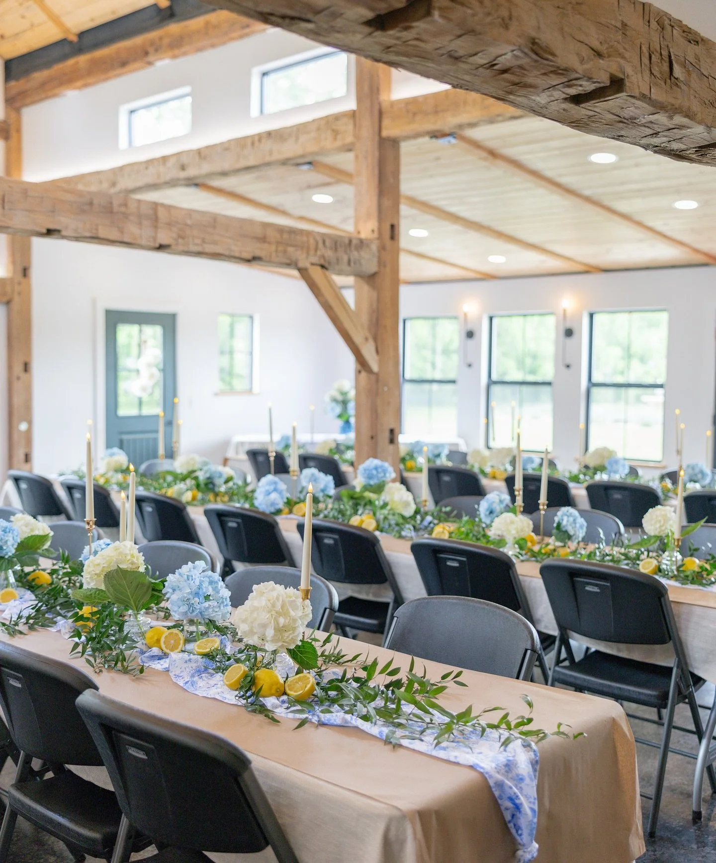 Interior of a rustic event space with wooden beams and large windows, decorated for a celebration with long tables covered in beige tablecloths, with floral centerpieces of white and blue hydrangeas, lemons, greenery, and tall white candles.