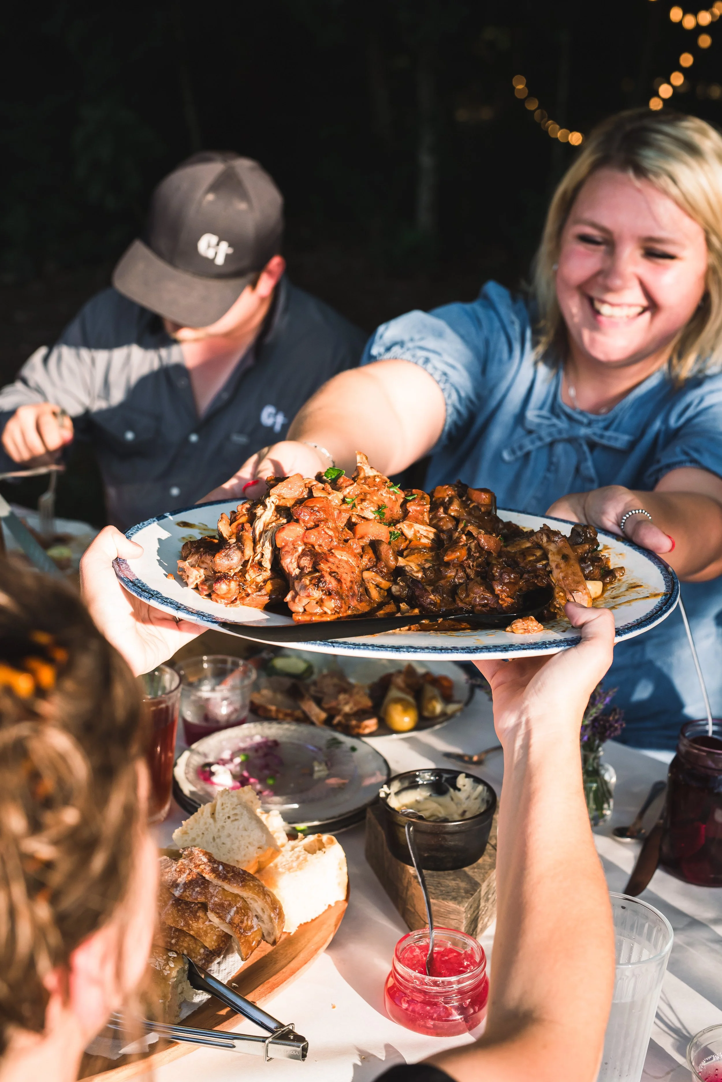 A woman in a blue shirt is smiling as she takes a plate of food from another person at The Farm to Table dinner party, with a table of food and drinks in front of her.