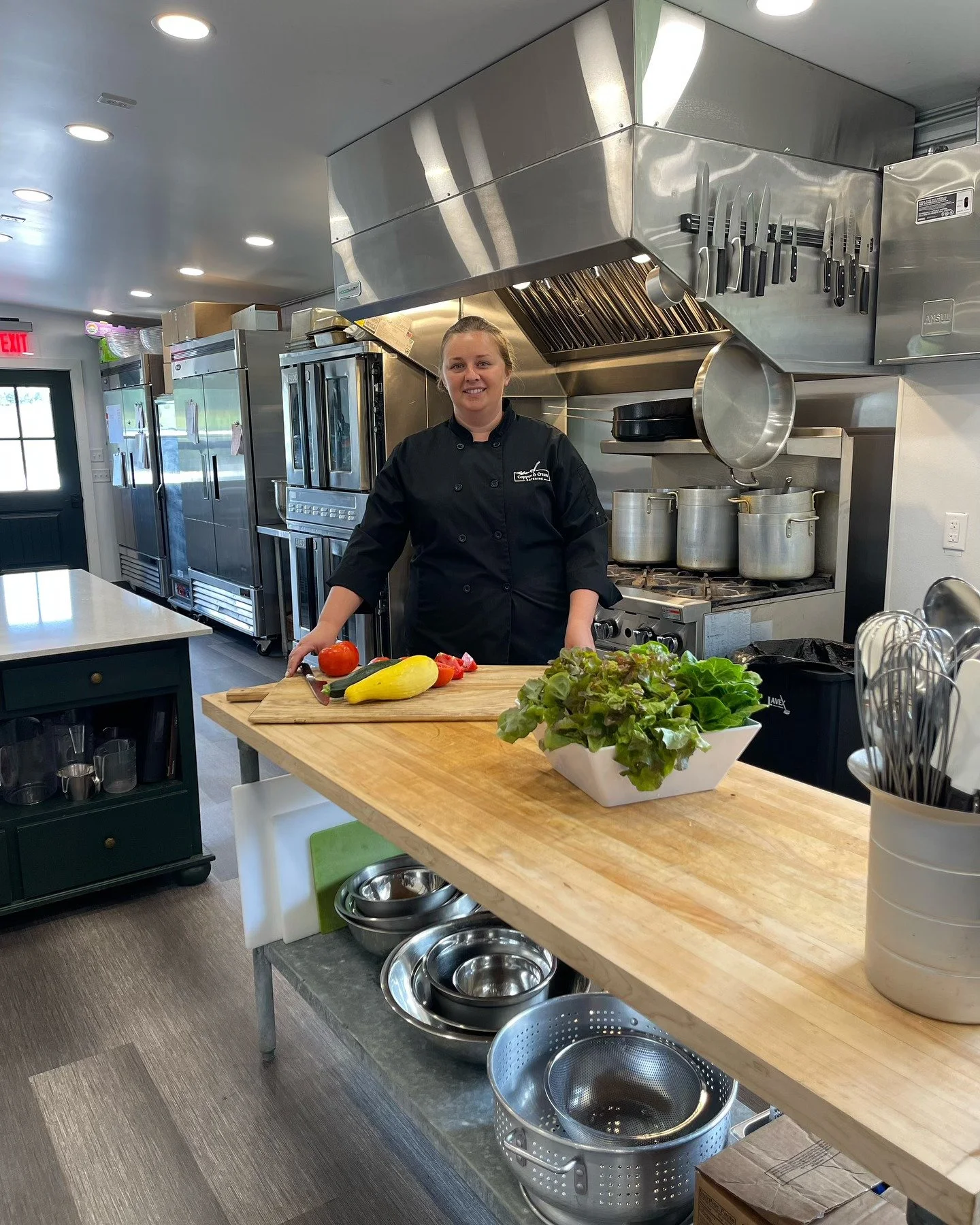 A female chef standing in a commercial kitchen, surrounded by kitchen equipment, with fresh vegetables in front of her on a wooden counter, smiling at the camera.