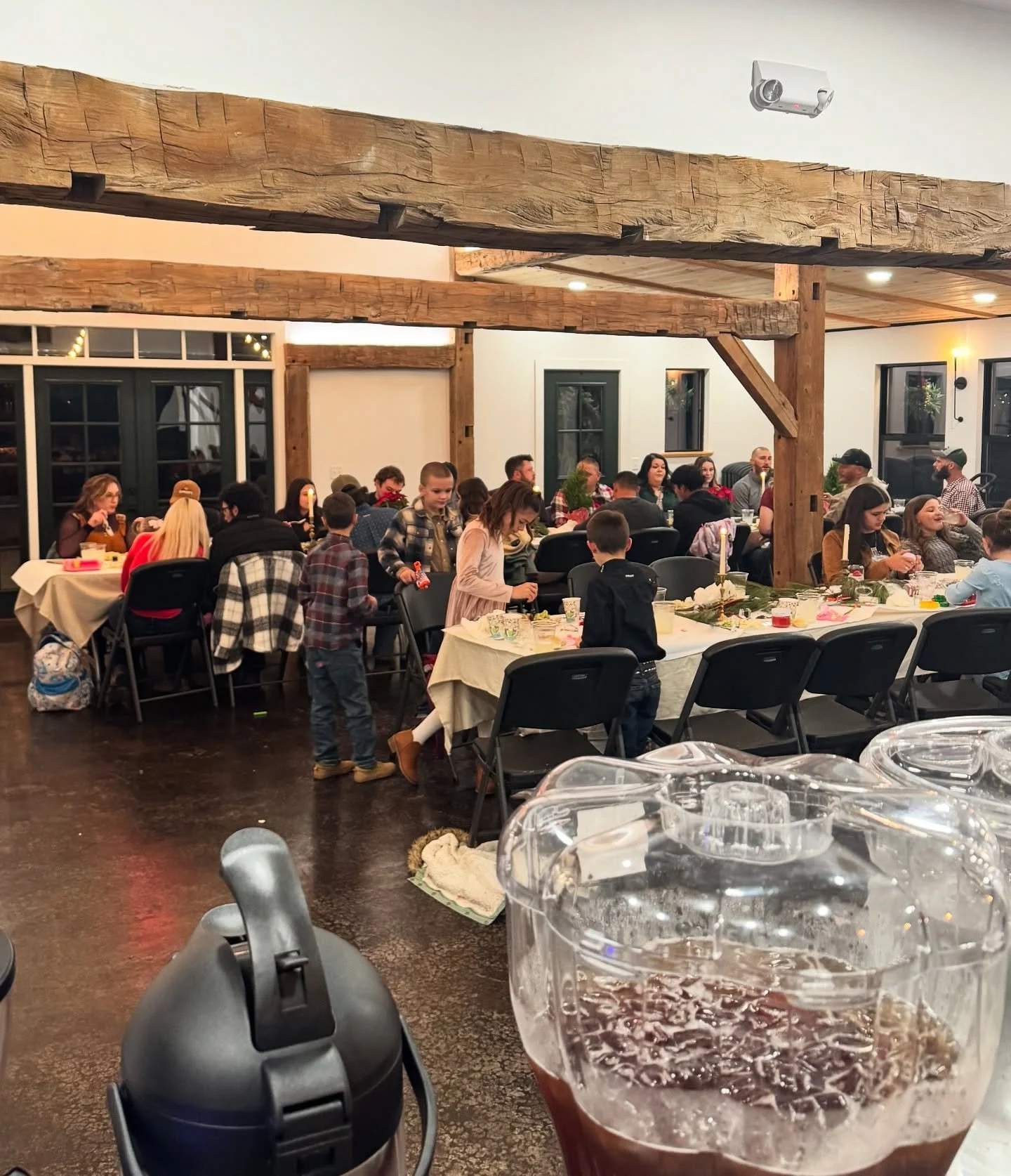 A large gathering of people, including children and adults, around several dining tables in a rustic-style restaurant or banquet hall with exposed wooden beams and a warm atmosphere, decorated with candles and holiday-themed items.
