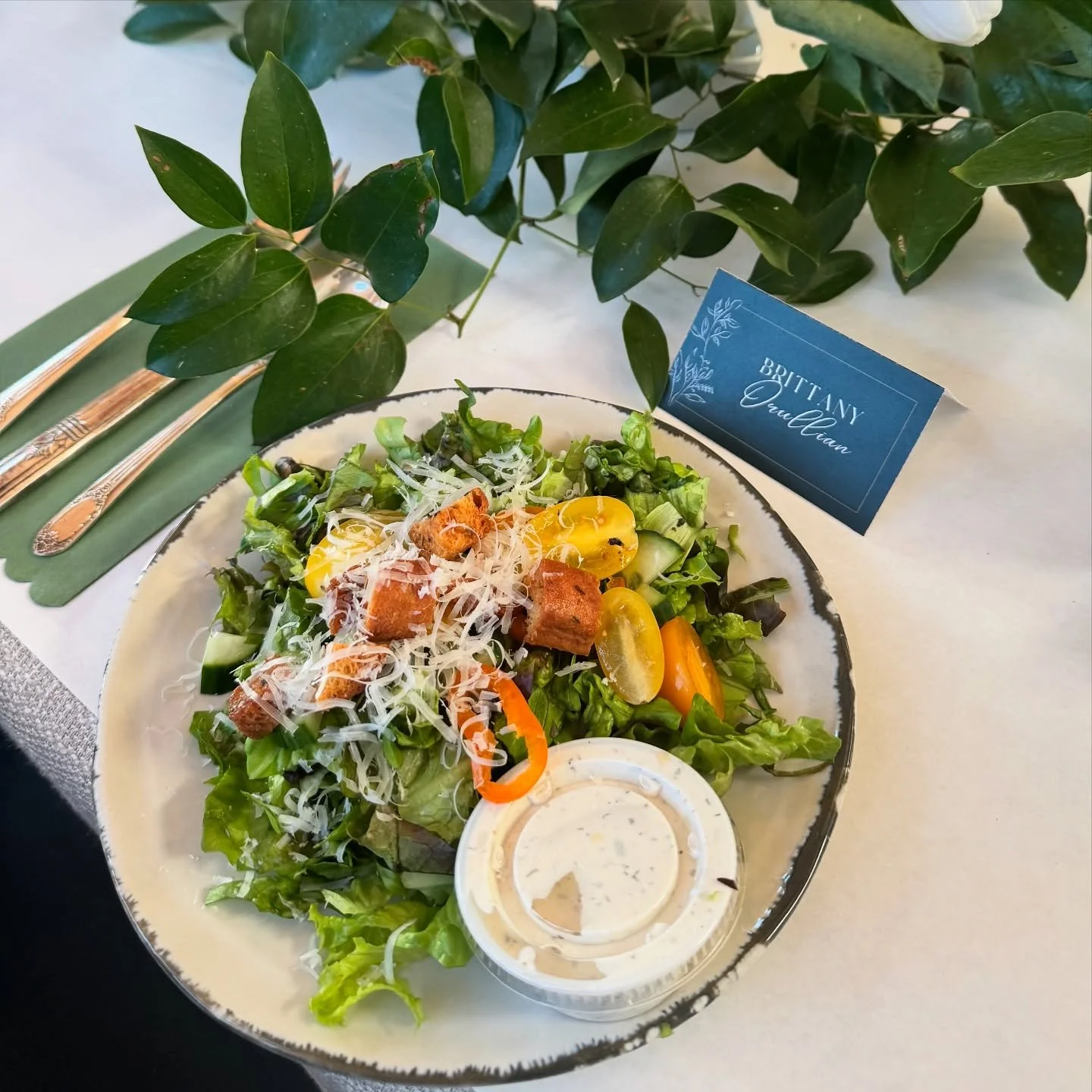A fresh mixed green salad with cherry tomatoes, shredded cheese, croutons, and a side of creamy dressing on a decorative plate, placed on a table with cutlery and a small name card that reads 'Britany Orellana,' surrounded by green foliage.