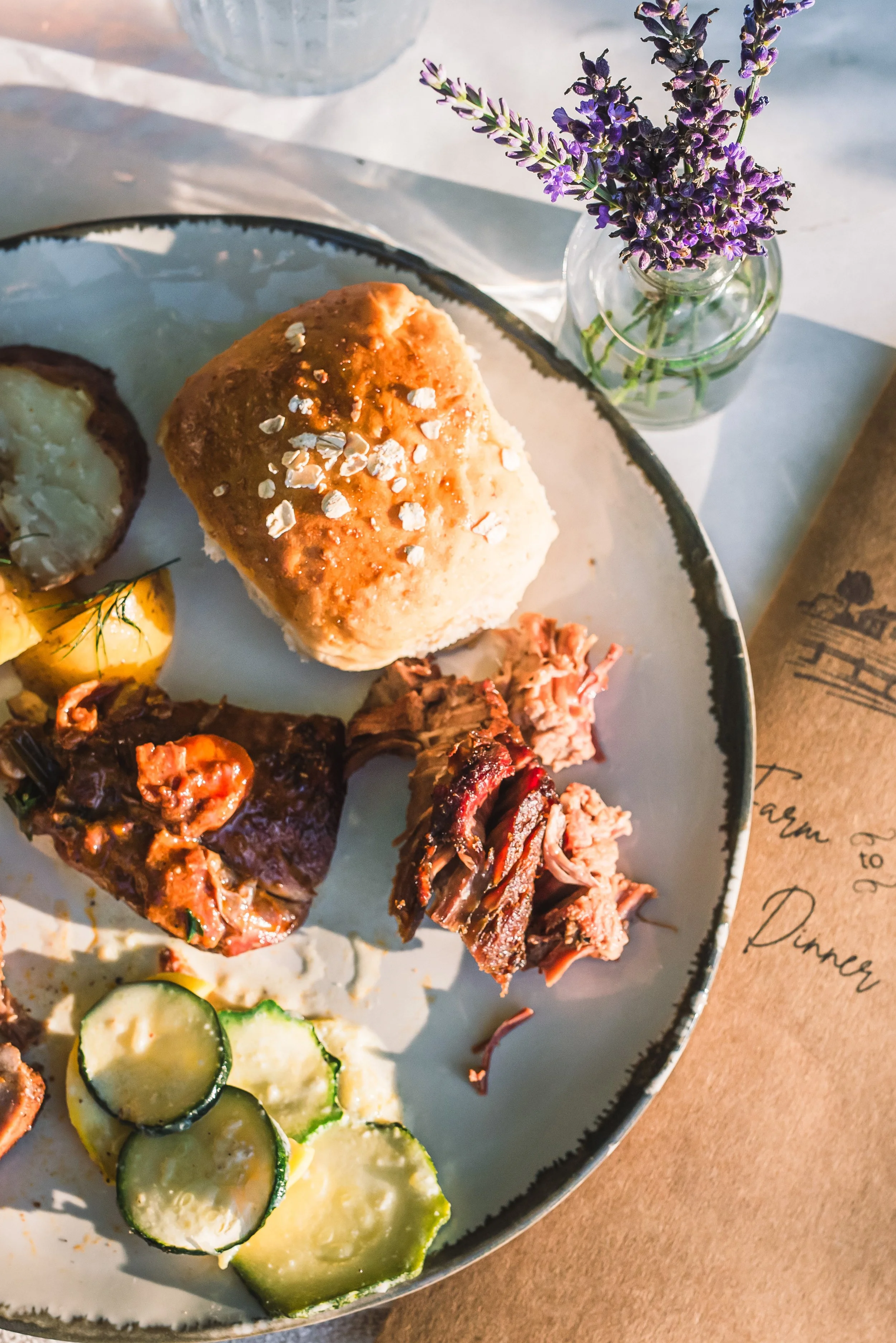 A plate of food with a roll of bread, slices of cucumber, a piece of grilled meat, shredded meat, and potatoes, accompanied by a small jar of purple flowers on a white table.
