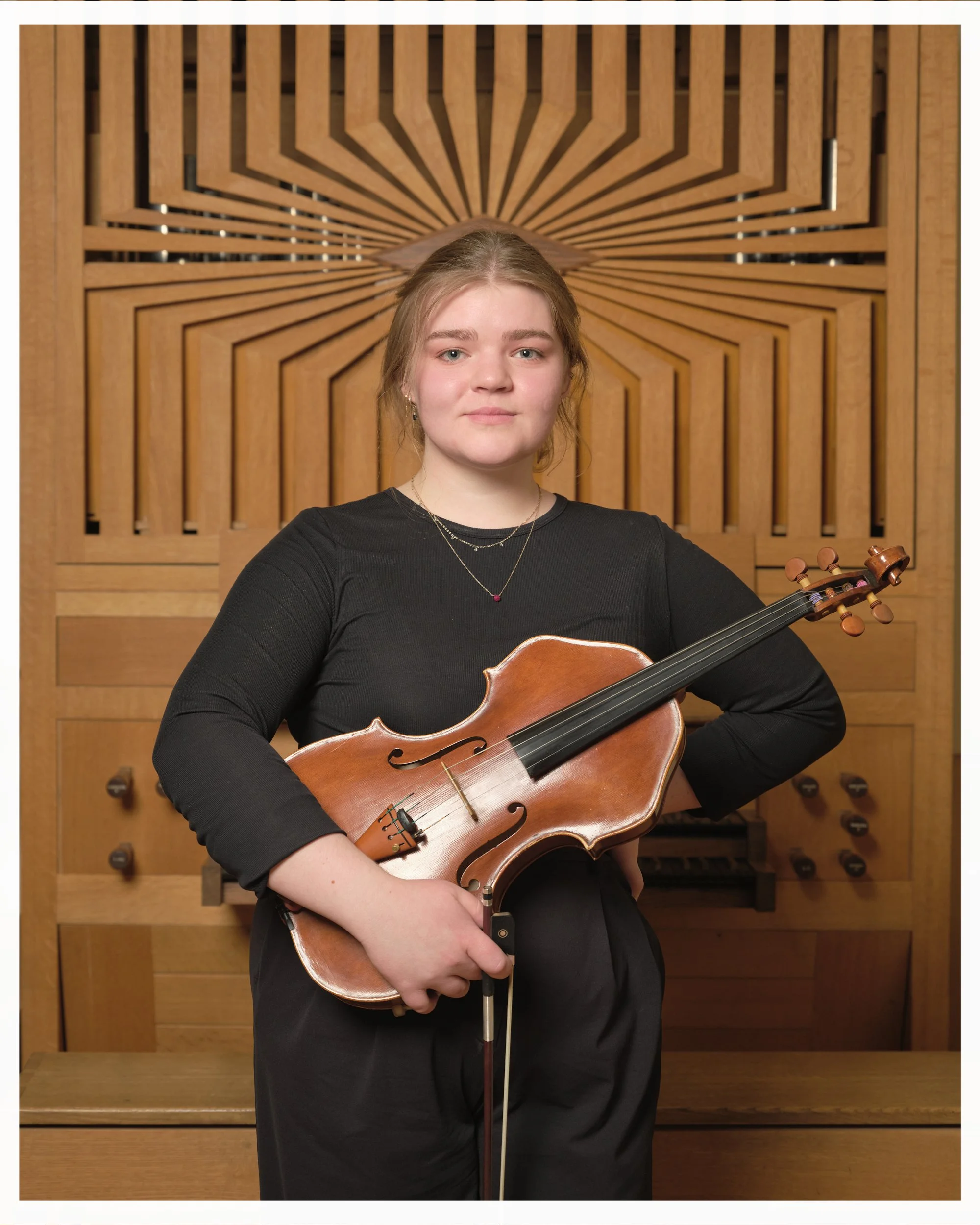 A young woman with light brown hair in a bun, wearing a black top and gold necklaces, holding a wooden violin in her left hand, standing in front of a wooden acoustic panel with geometric patterns.