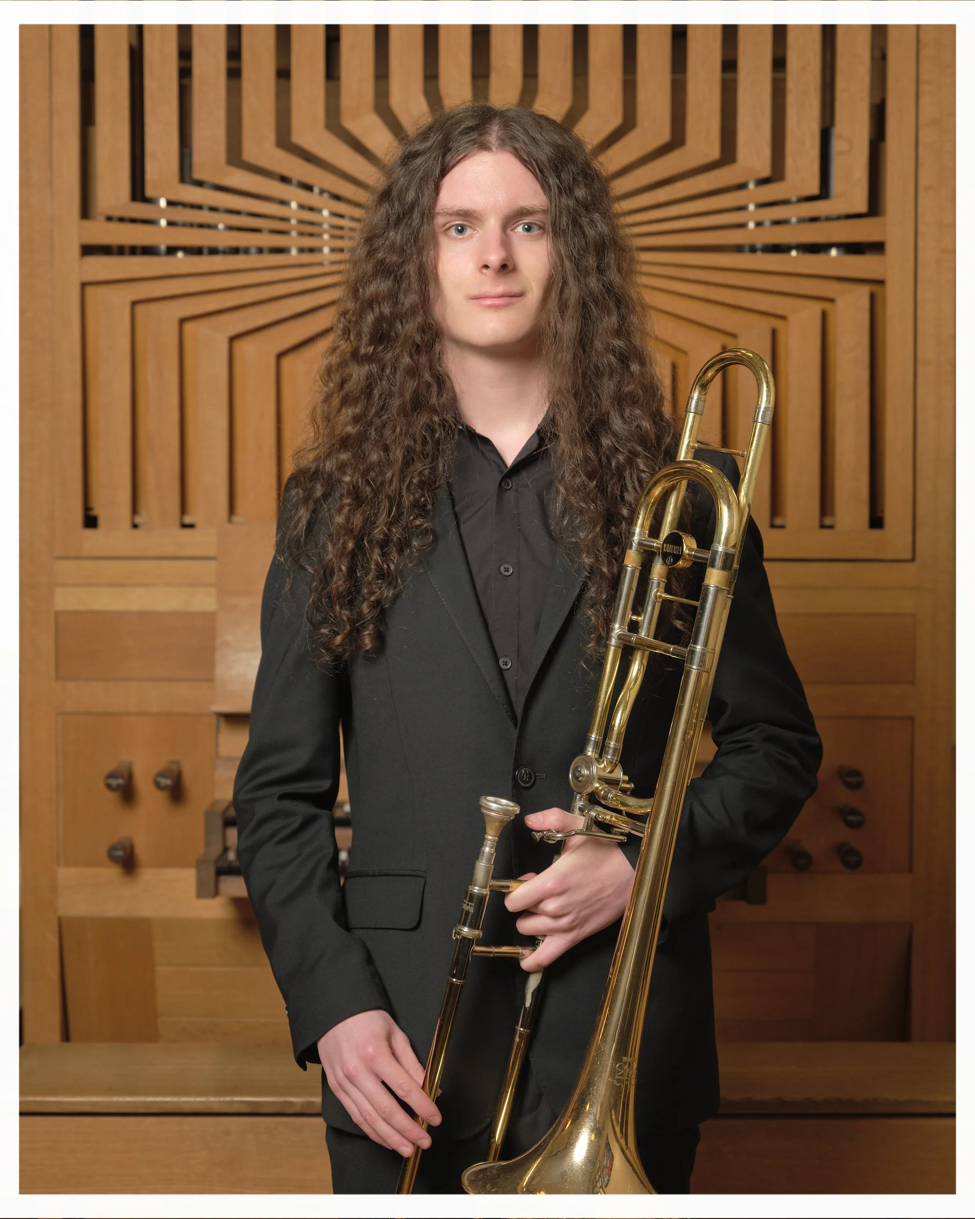 A young man with long curly hair wearing a black suit holding a trombone, standing in front of a wooden acoustic panel.