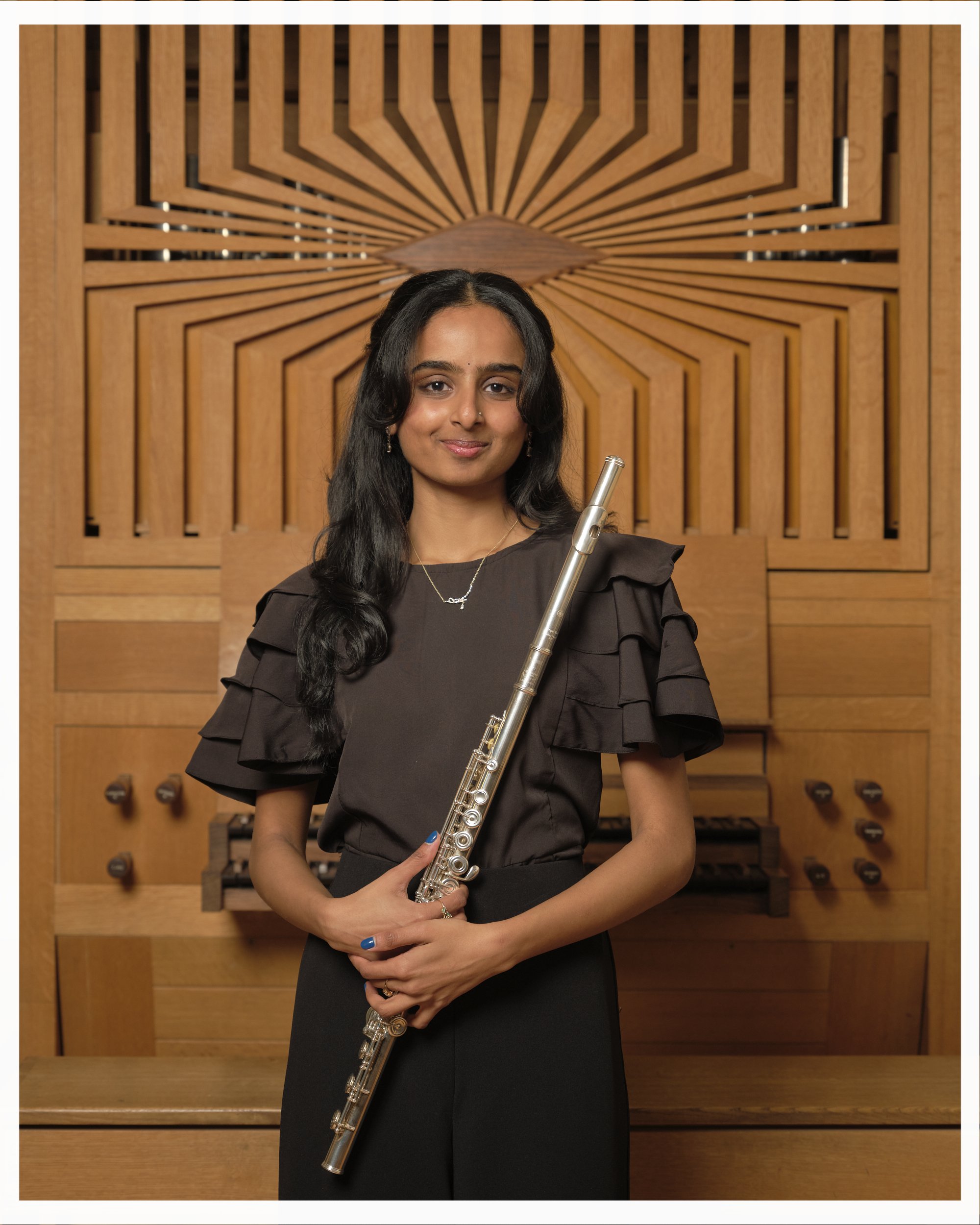 A woman with long black hair holding a flute, standing in front of a wooden wall with decorative geometric patterns.