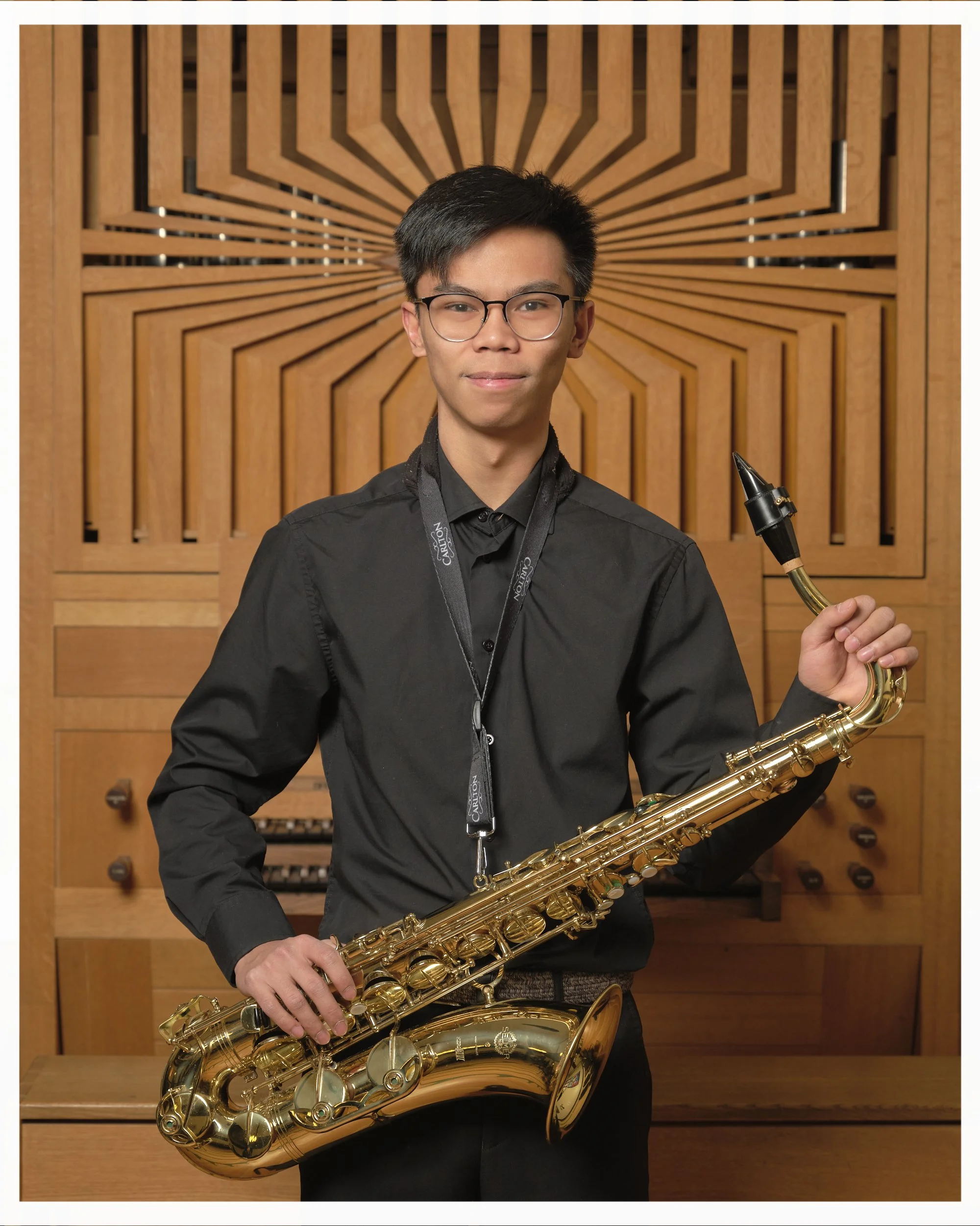 Young man standing in front of a wooden panel, holding a gold-colored alto saxophone, wearing glasses, a black shirt, and a lanyard with the word 'Canton'.