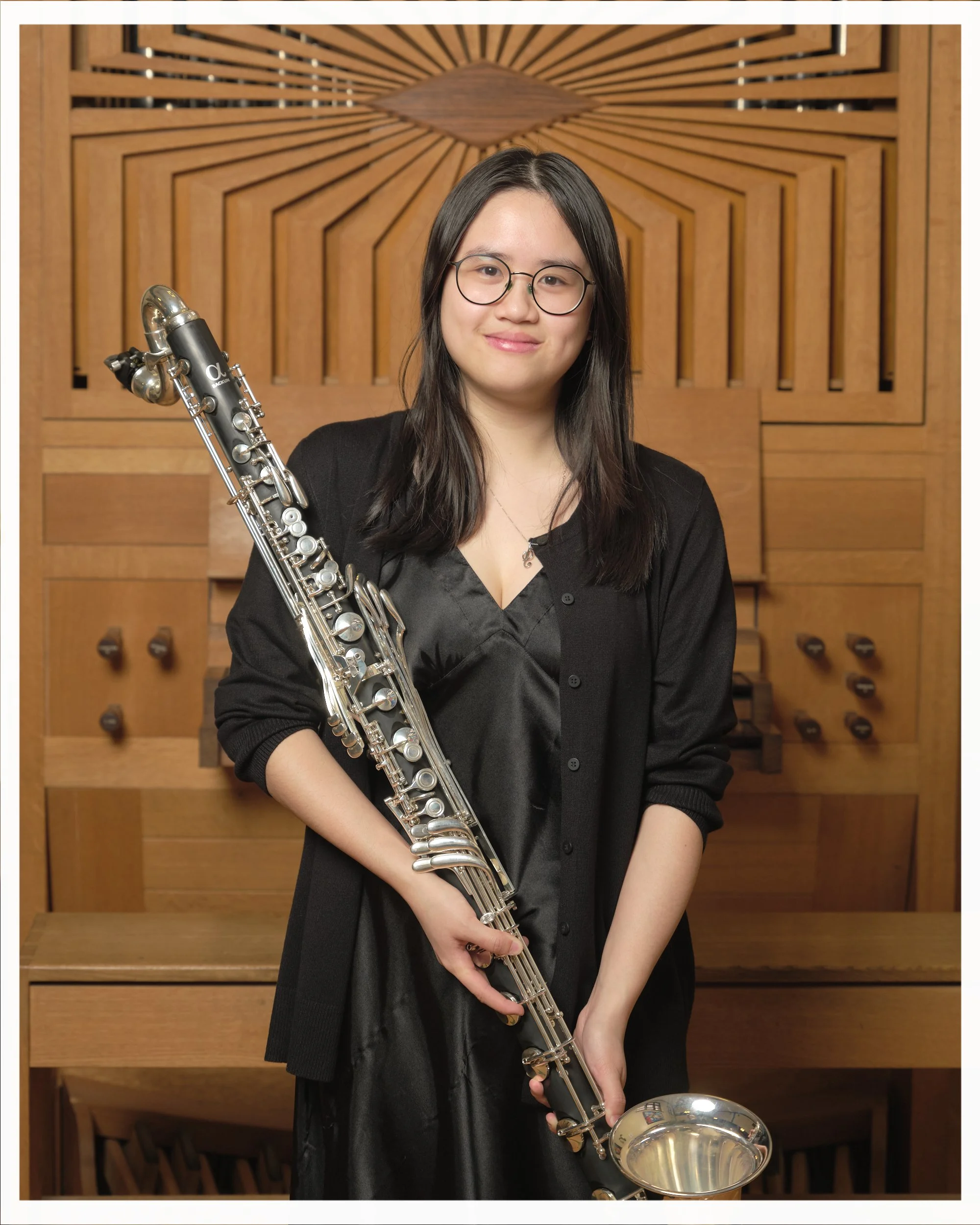 A young woman with glasses standing in front of a wooden pipe organ, holding a silver soprano saxophone, wearing a black dress and cardigan.
