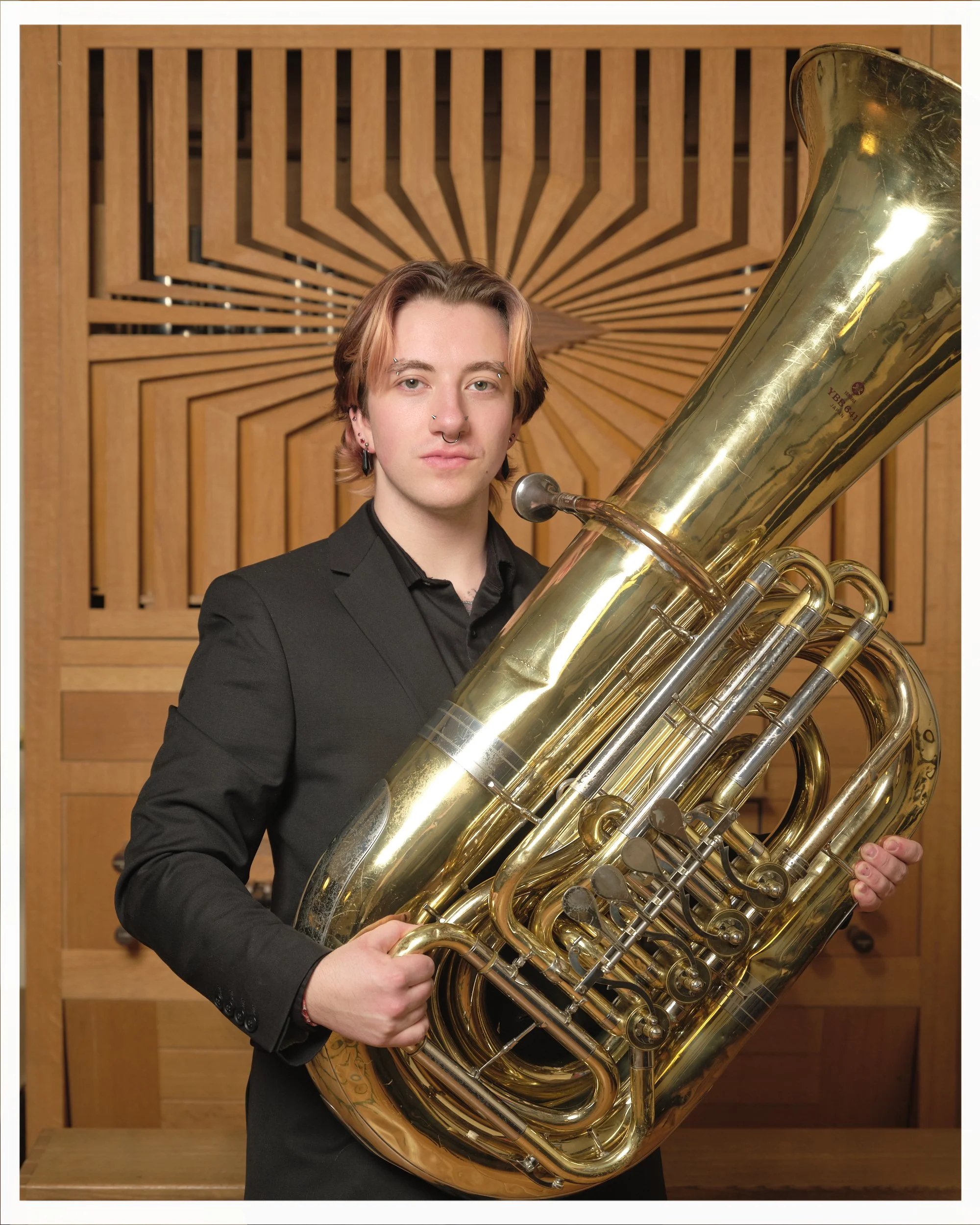 A young person with medium-length hair, wearing a black suit, holding a large brass tuba in front of a wooden decorative wall.