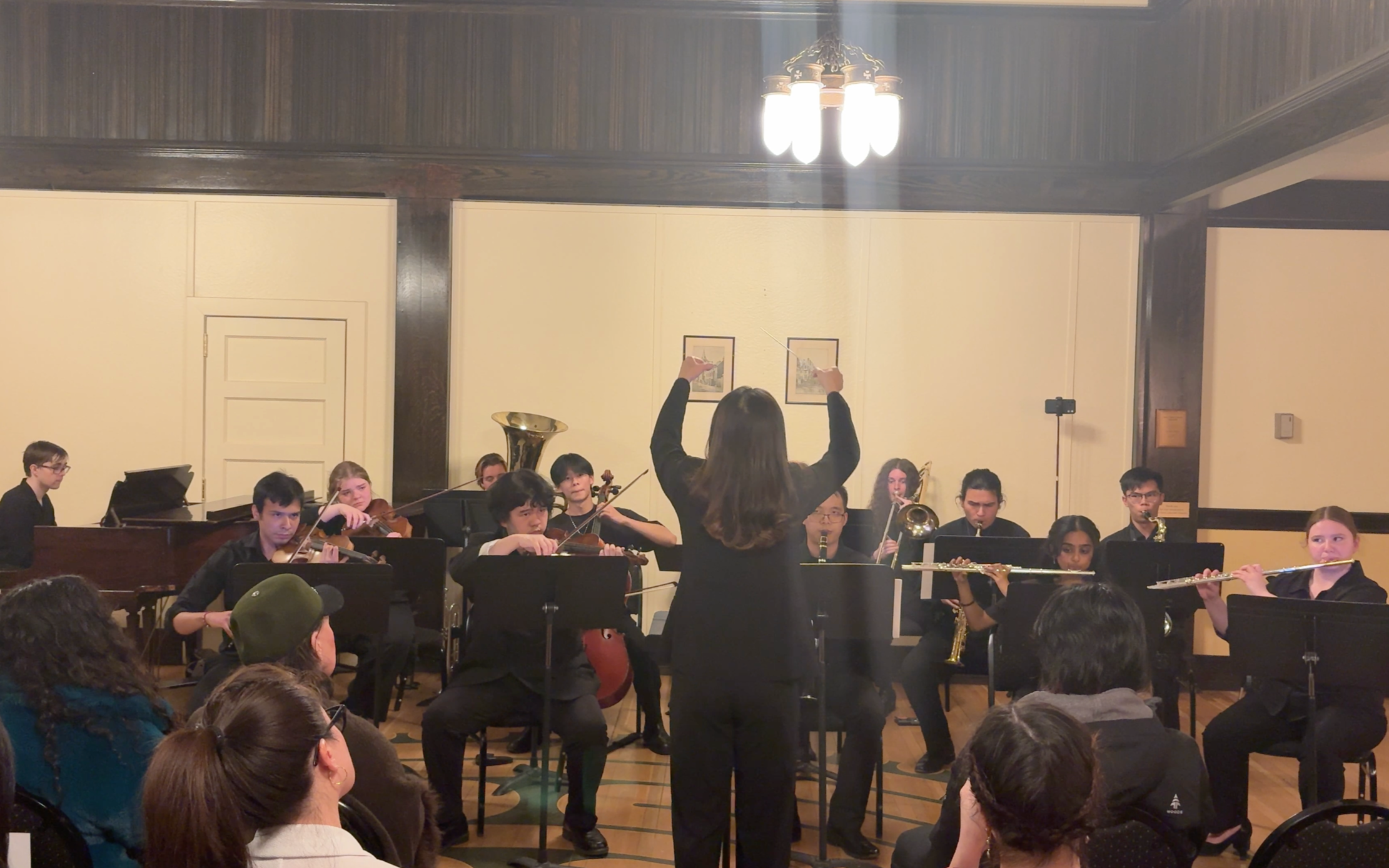 Orchestra performing on stage with a conductor leading, audience seated in front, in a room with beige walls and wooden accents.