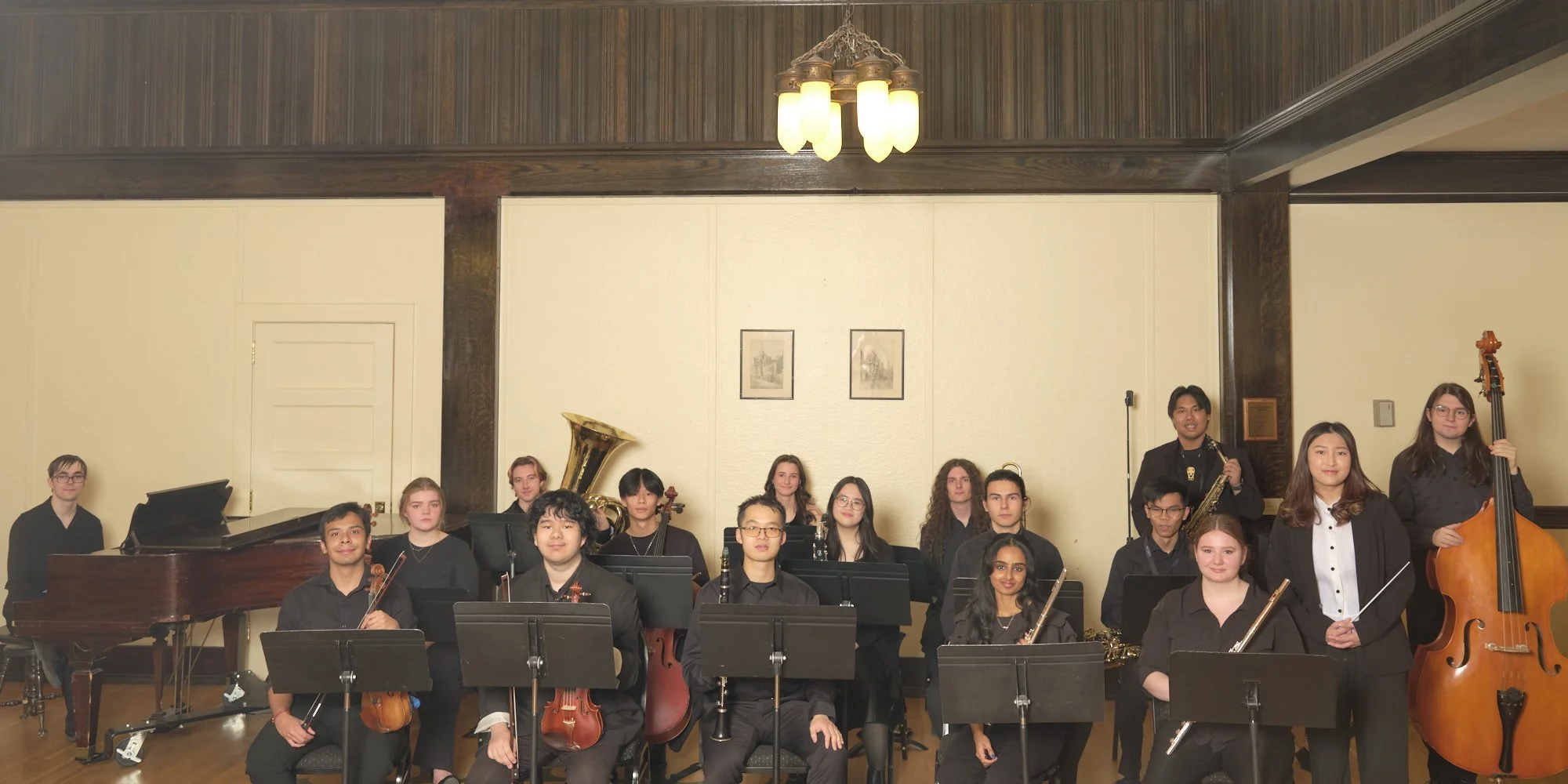 A group of young musicians in black attire holding various musical instruments in a room with cream walls and wood paneling, posing for a photo.