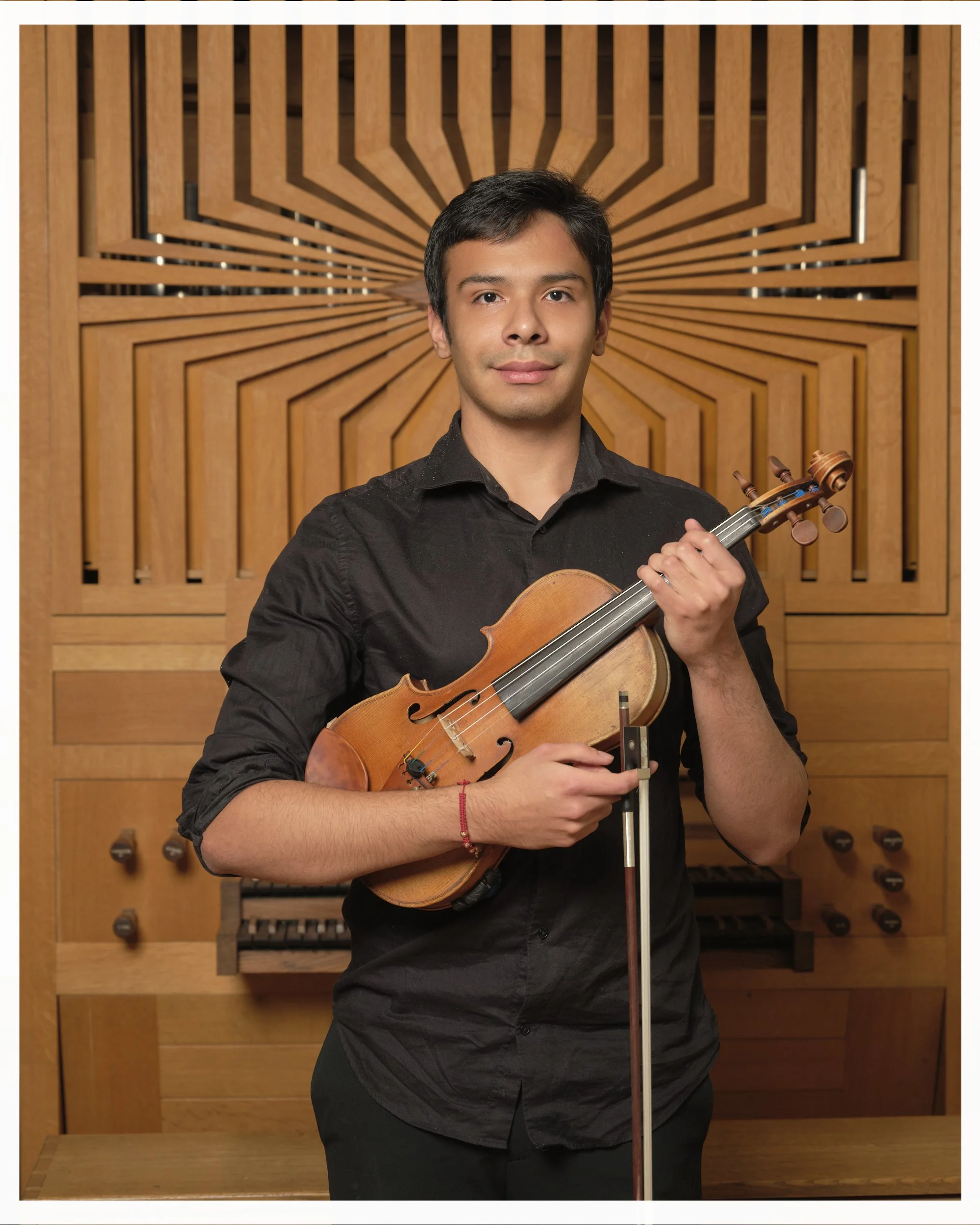 A young man holding a violin and bow in a music studio with wooden acoustic panels and organ pipes in the background.