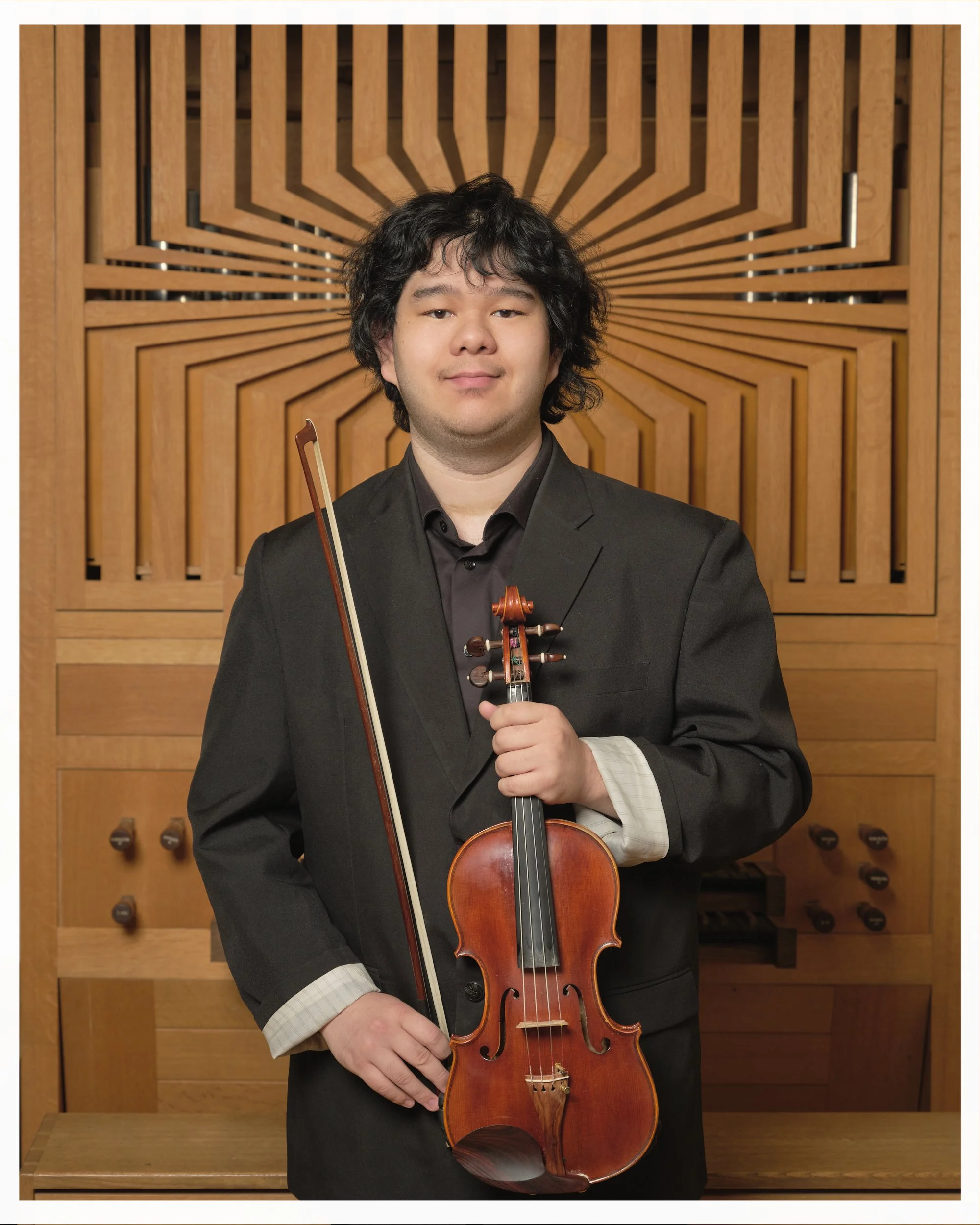 A young man in a black suit holding a violin and bow, standing in front of an acoustic panel background.
