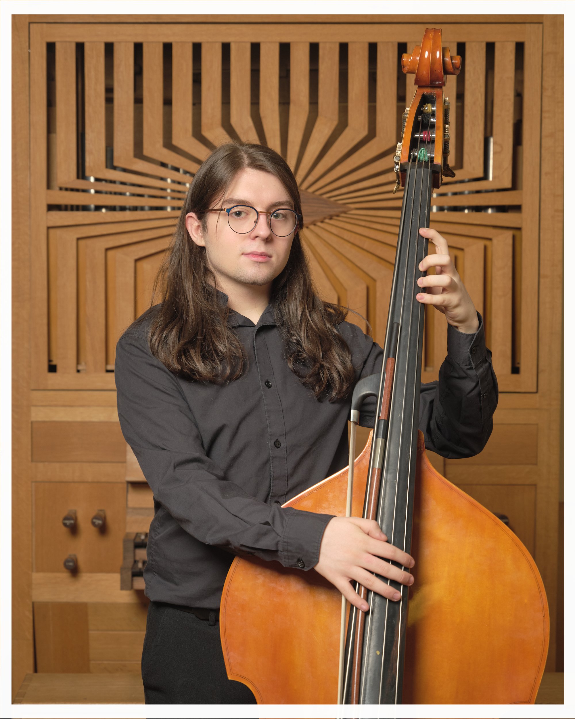 A young man with long brown hair, glasses, and a black shirt, playing a double bass in a music room with wooden acoustic panels.