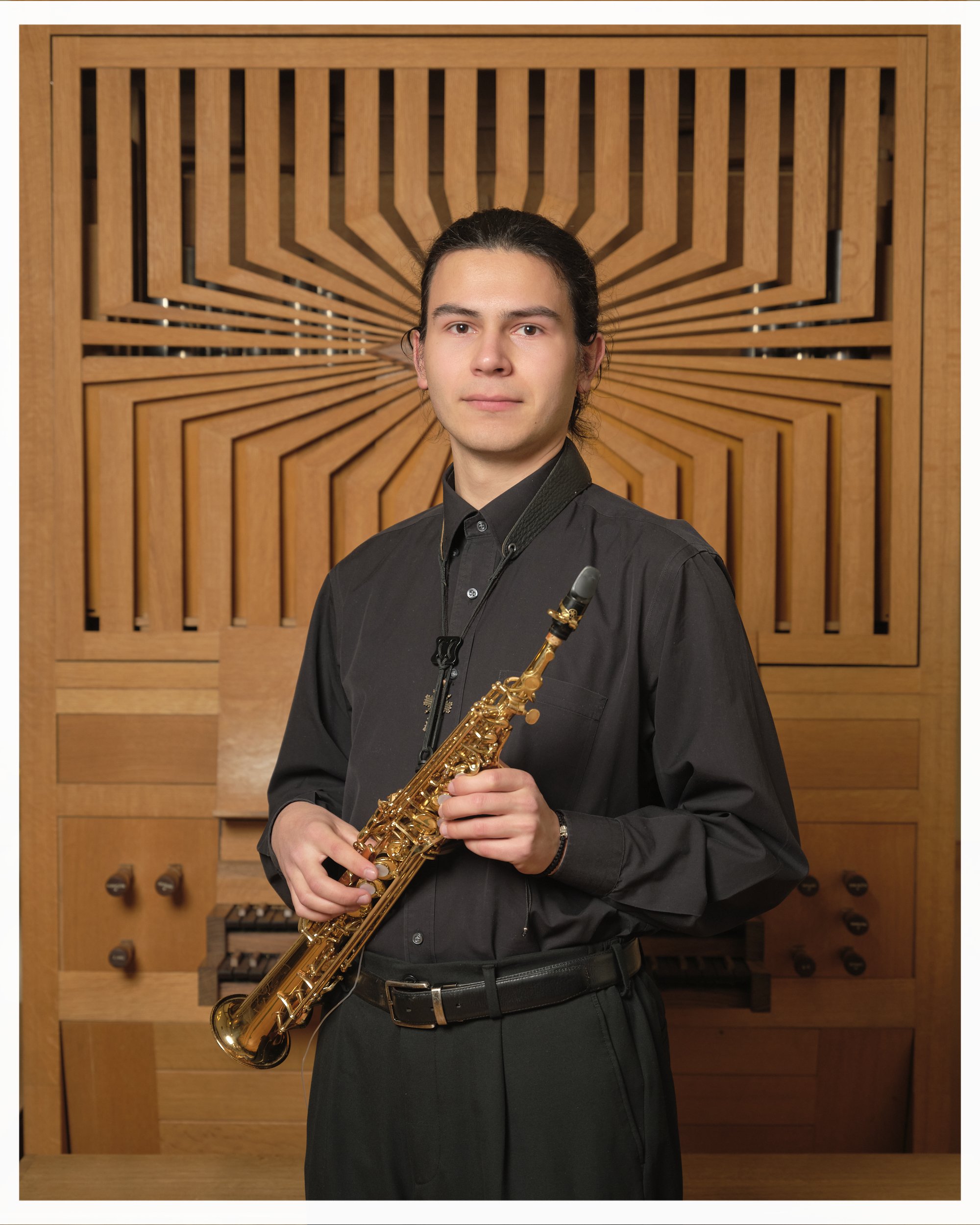 A young man with long dark hair, wearing a black shirt, holding a gold-colored saxophone, standing in front of a wooden background with geometric patterns.