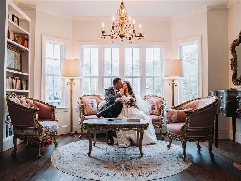 Bride and groom kissing on a sofa in an elegant living room with chandelier and tall windows.