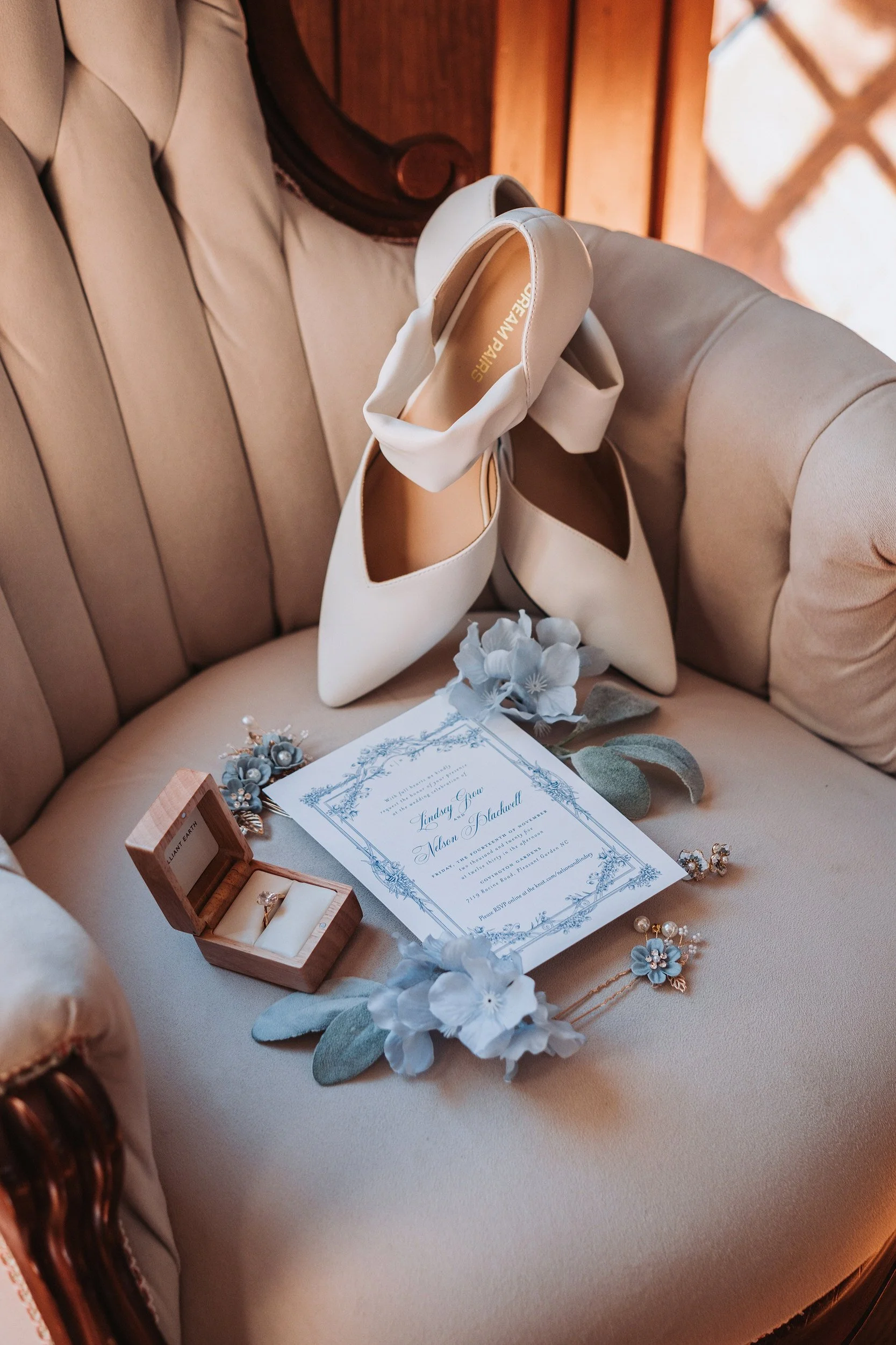A pair of white high heels, a wedding invitation, a small wood jewelry box with an engagement ring, blue flowers, and bridal accessories arranged on a vintage beige couch.