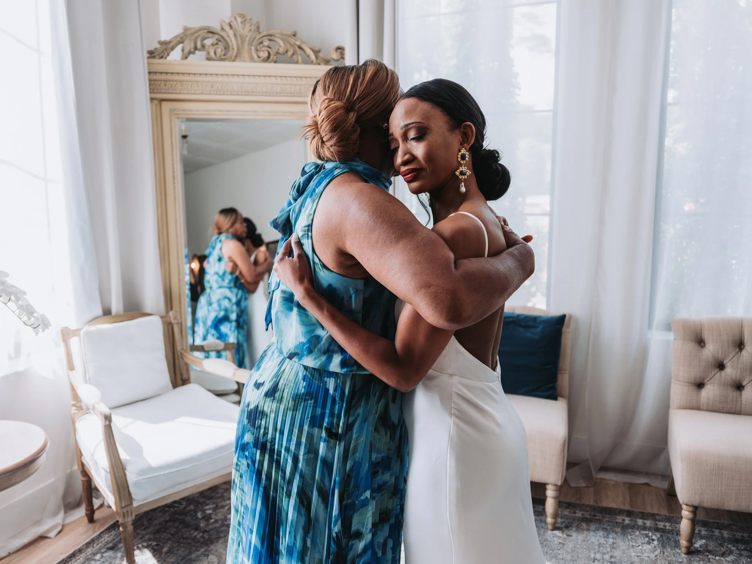 Two women embracing in a softly lit room, with one wearing a white dress and the other in a blue patterned dress, and a mirror reflecting their hug.
