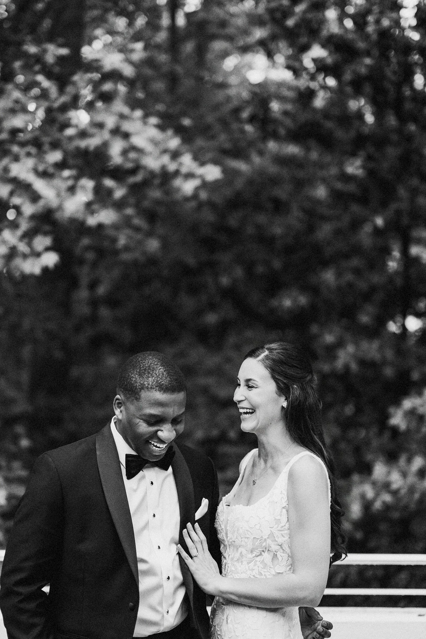 A joyful couple, dressed in wedding attire, laughing and smiling outdoors with trees in the background.