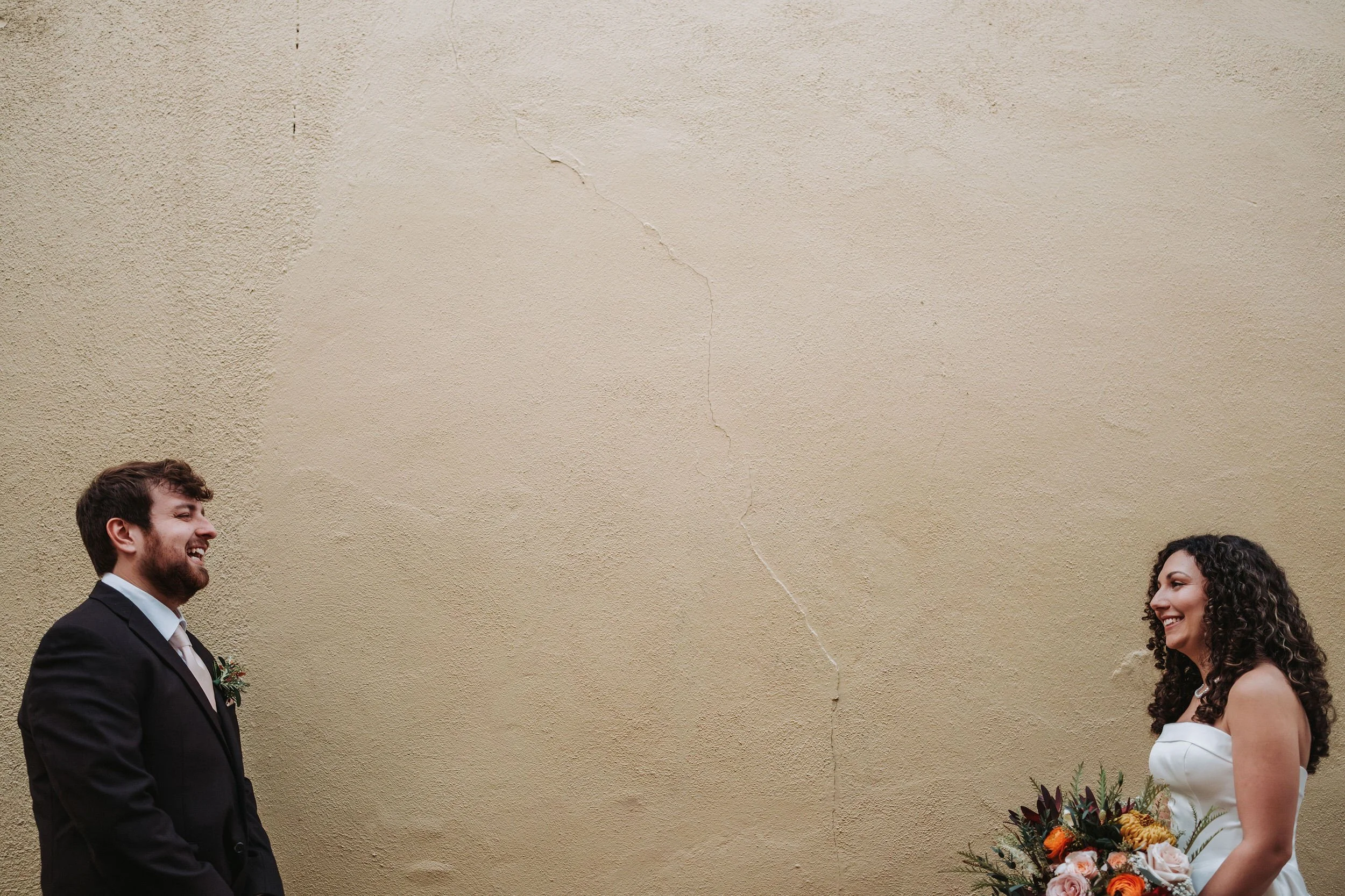 A bride and groom standing apart facing each other, smiling, on a beige wall background with a crack.