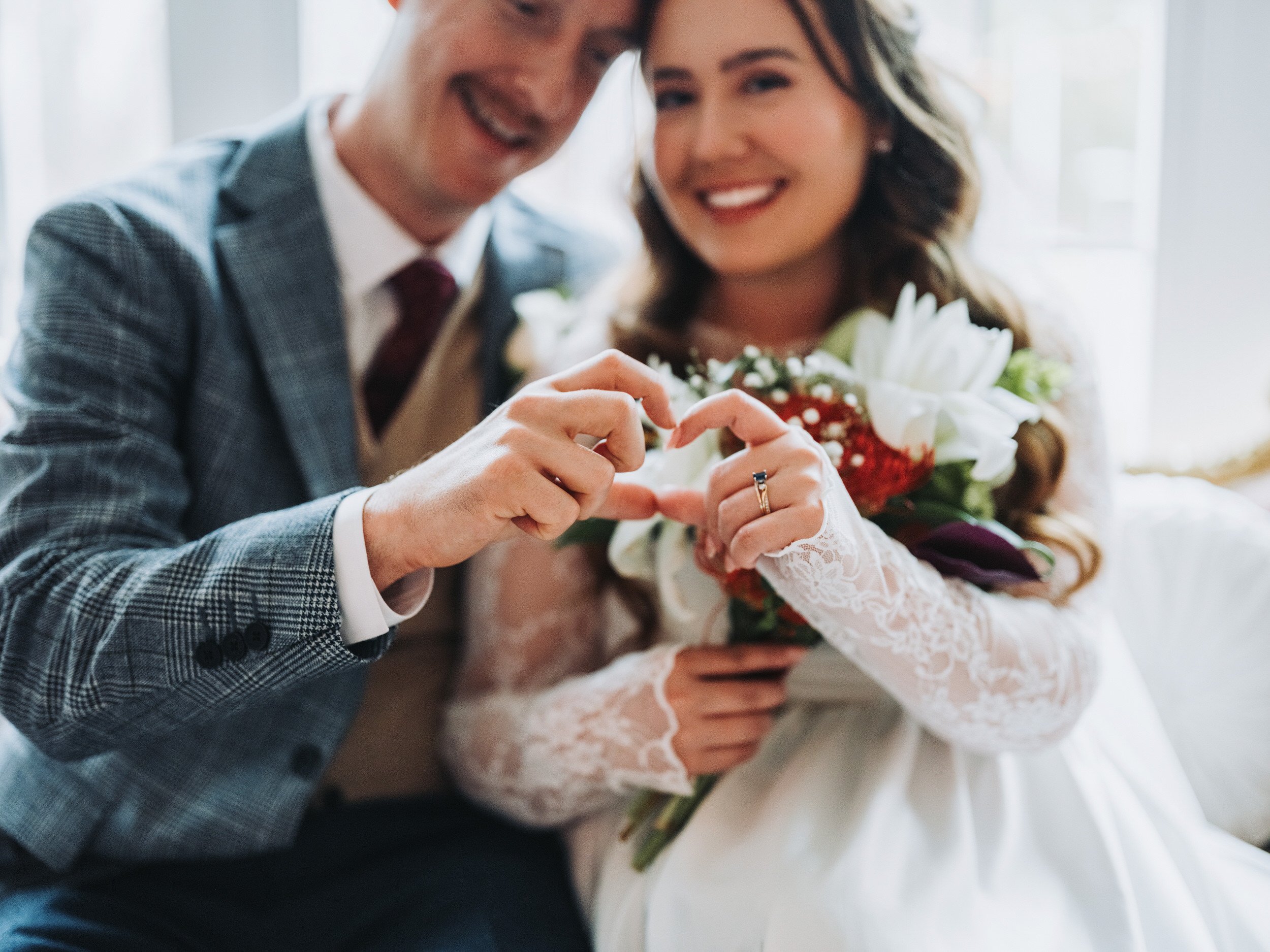 A newlywed couple smiling and forming a heart shape with their hands, with the bride holding a bouquet of flowers.