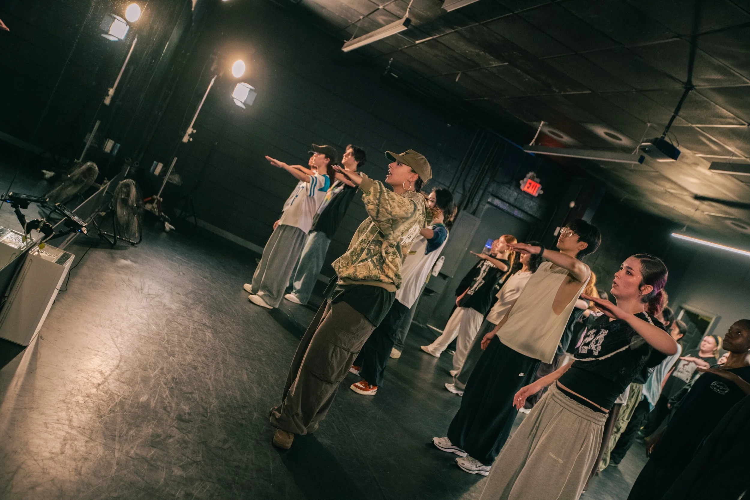 Group of diverse people standing in a dance or fitness studio, practicing a synchronized movement with one arm extended forward in a line formation.