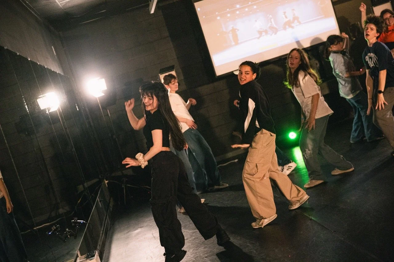 A group of young people dancing together in a dimly lit room with a large screen displaying a lighted scene in the background.