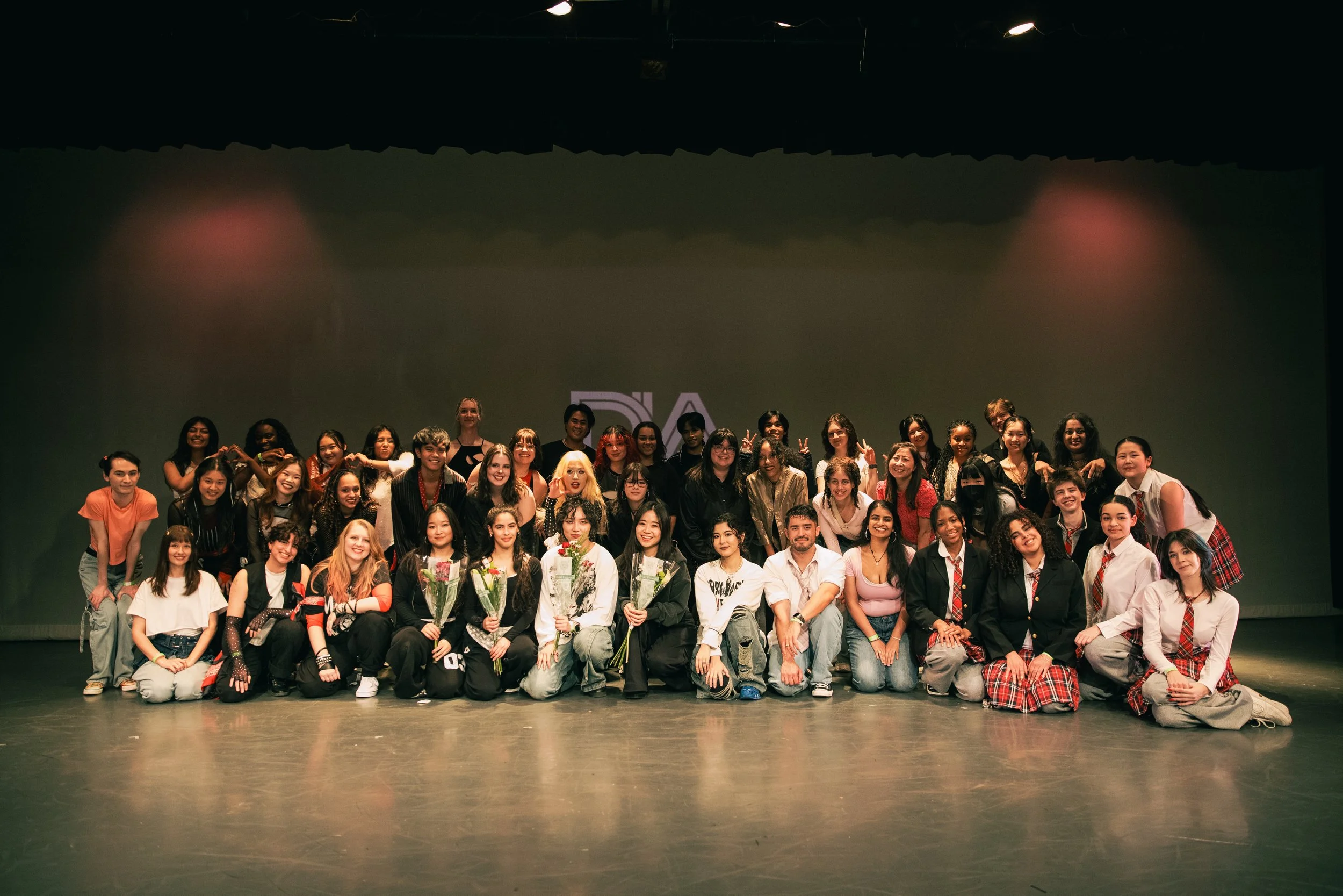 Group of people on a stage, posing for a photo after an event or performance, with some holding flowers and making peace signs.