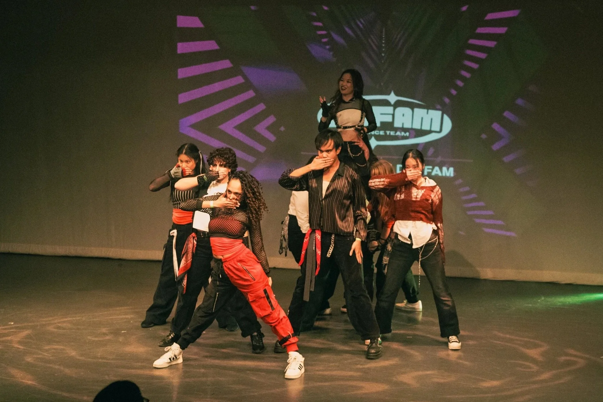 Group of seven dancers on stage performing in front of a purple and blue geometric background with the logo 'FAM' visible.