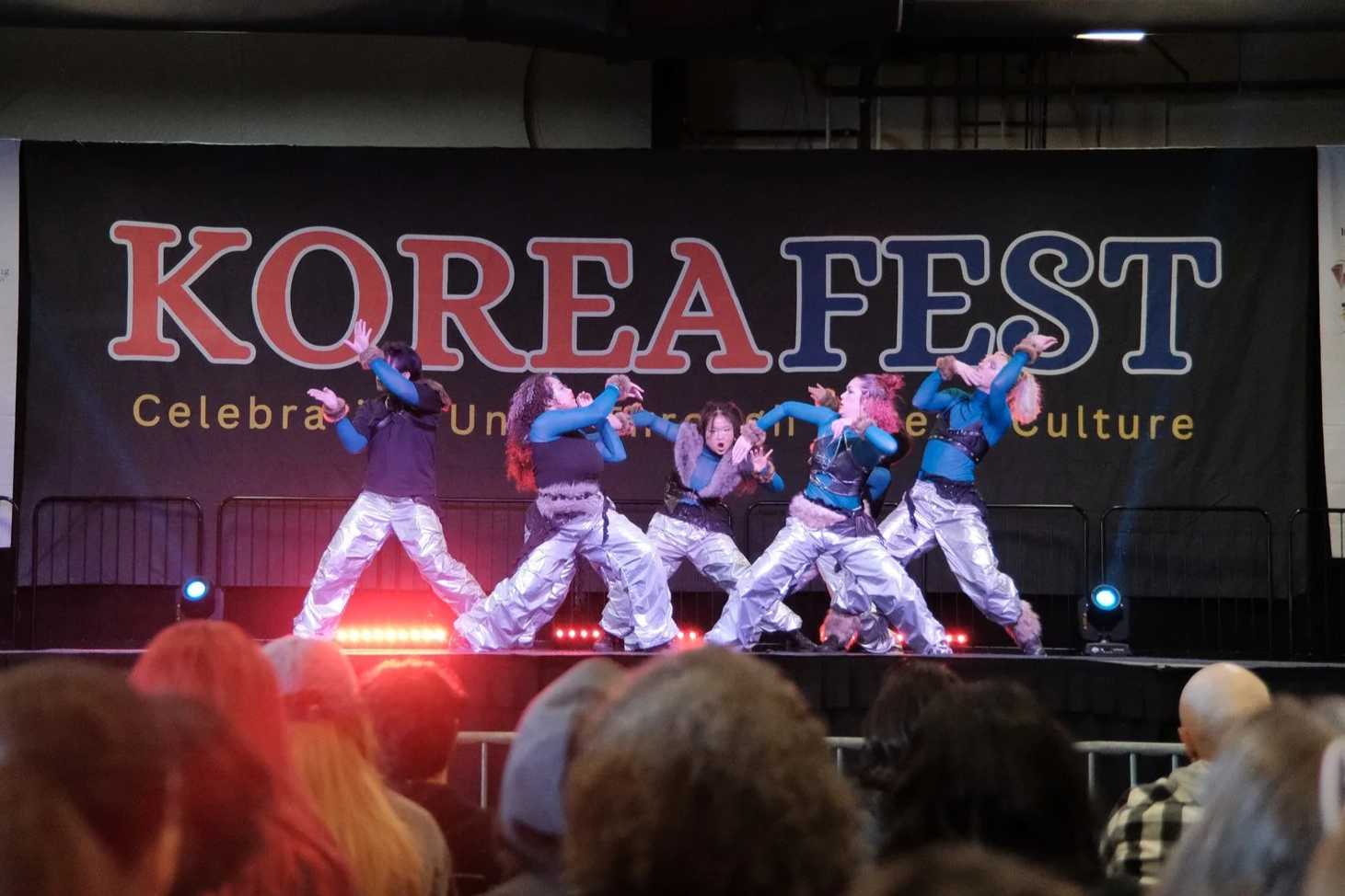 A group of six performers on a stage at Korea Fest, dancing in coordinated outfits with metallic pants and blue sleeves, with an audience watching.