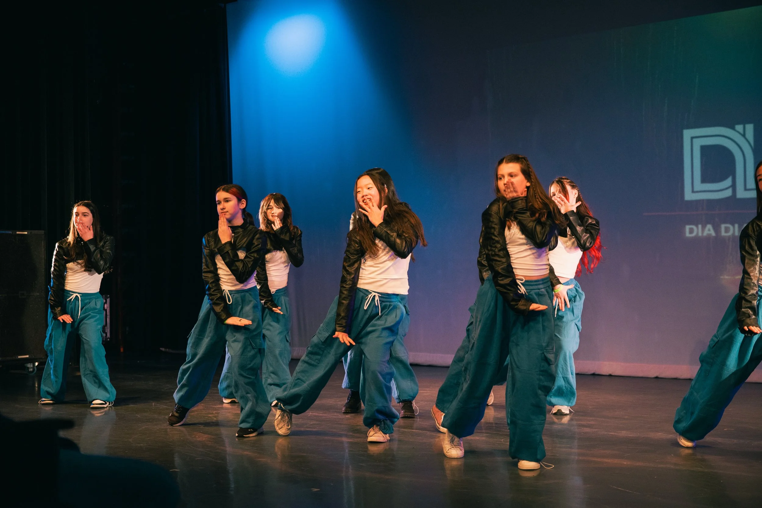 Group of young girls performing a dance on stage, wearing matching teal pants, black and white jackets, and sneakers, with some covering their mouths and others smiling, under stage lighting.