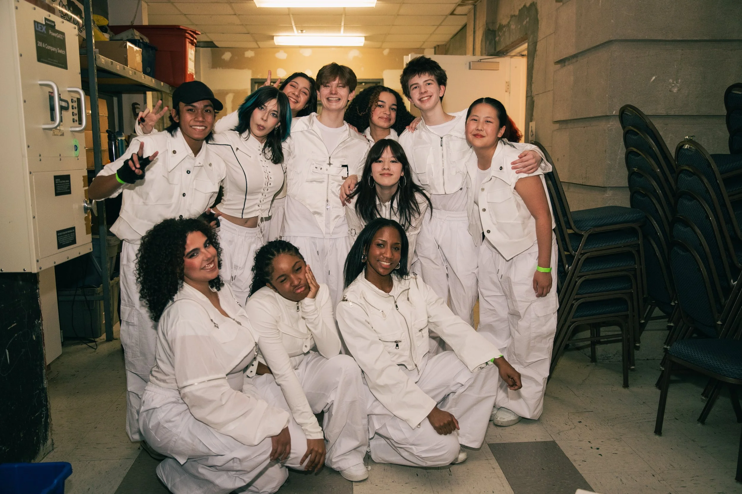 A group of young people in white jumpsuits posing and smiling for a photo in an indoor setting that appears to be a storage room or basement.