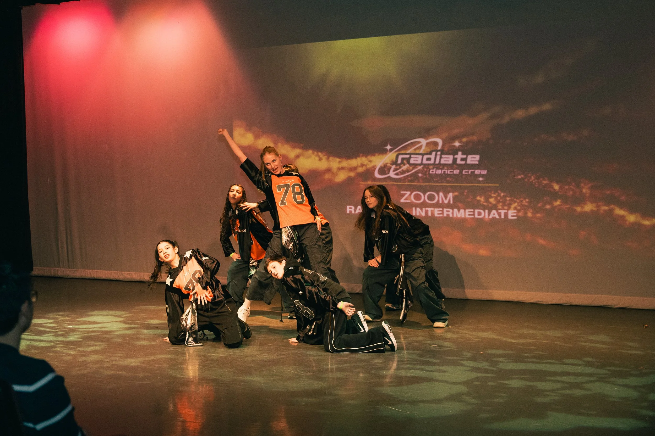 Group of young dancers performing on stage, some kneeling and others standing, with a colorful backdrop displaying the words 'Radiate Dance Crew,' 'Zoom,' and 'Intermediate'.