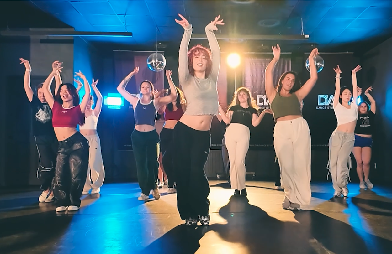 Group of diverse young women posing in a dance studio with black walls, red lighting, and 'DIA Dance Studio' posters behind them, some making finger guns and smiling.