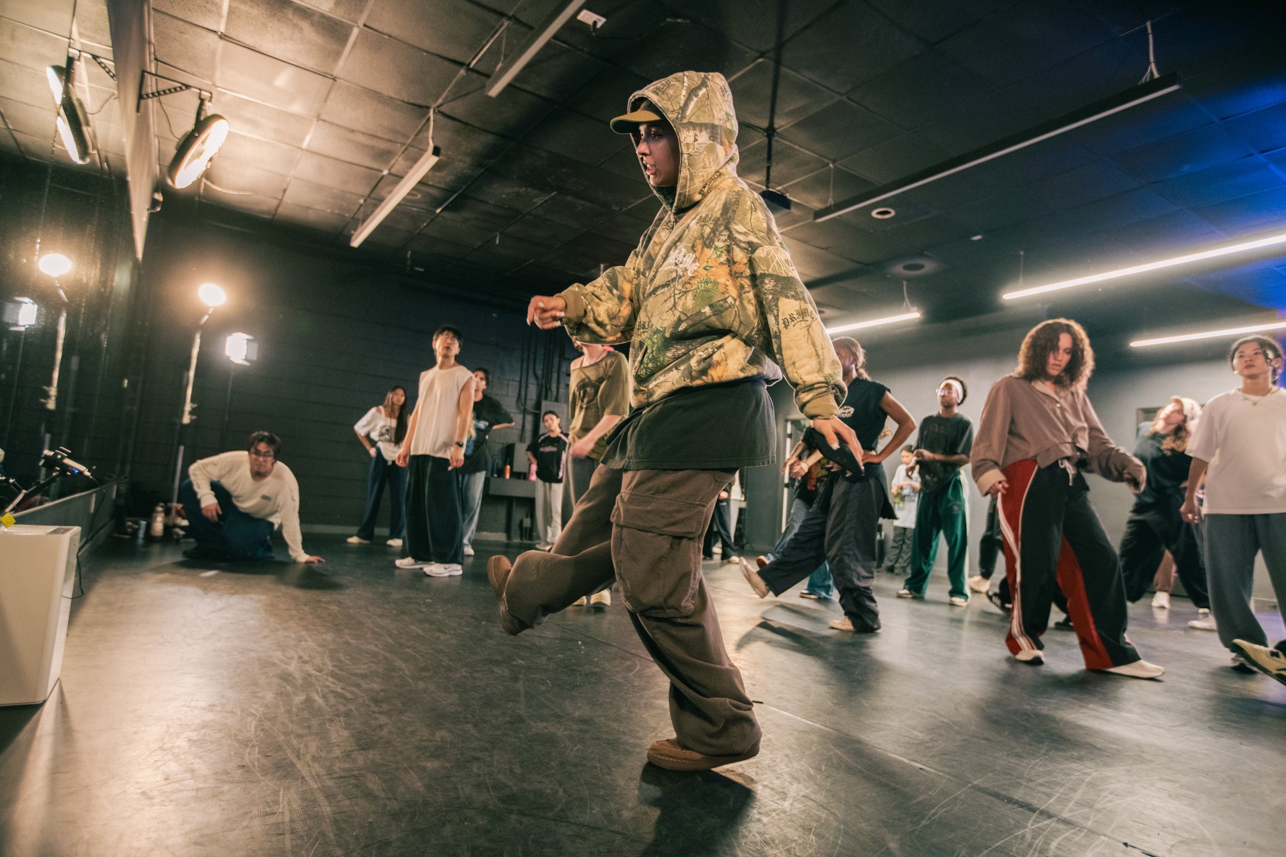 Group of people dancing and rehearsing in a dance studio with black walls and ceiling, illuminated by overhead lights. The individuals are dressed casually, with some wearing hoodies and sweatpants.