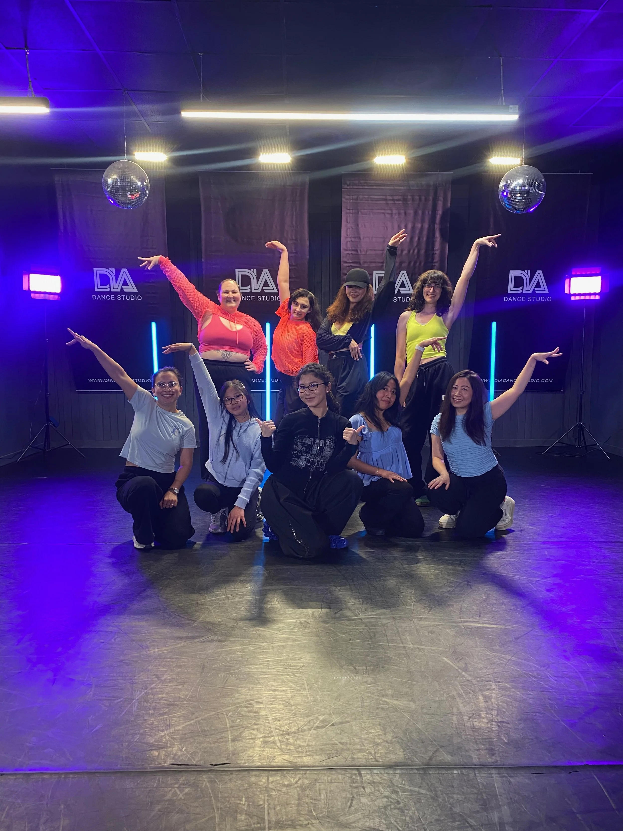 Group of ten women standing in a dance studio with black walls, illuminated by purple and blue lights, with two disco balls hanging from the ceiling and dance studio banners in the background. The women are posing with raised arms, smiling, dressed in casual active wear.
