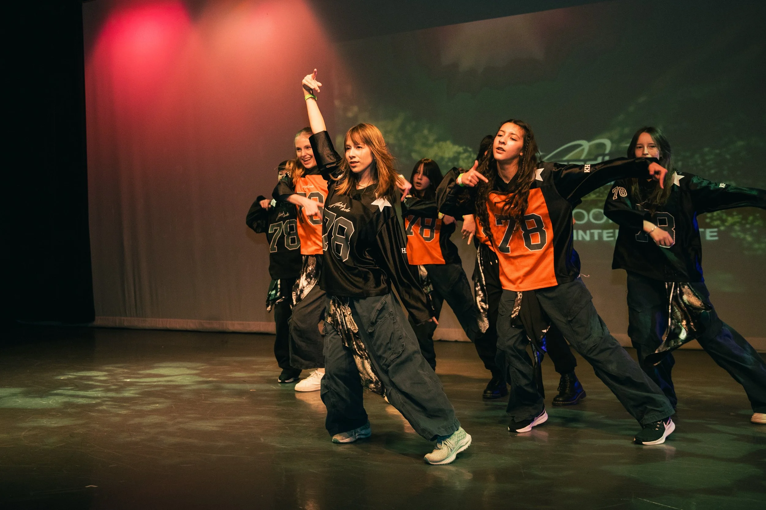 A group of young women dancing on stage during a performance. They are dressed in coordinating black and orange outfits, with some wearing oversized black jackets and baggy pants. The background is dark with a digital display or projection, and stage lights illuminate the scene.