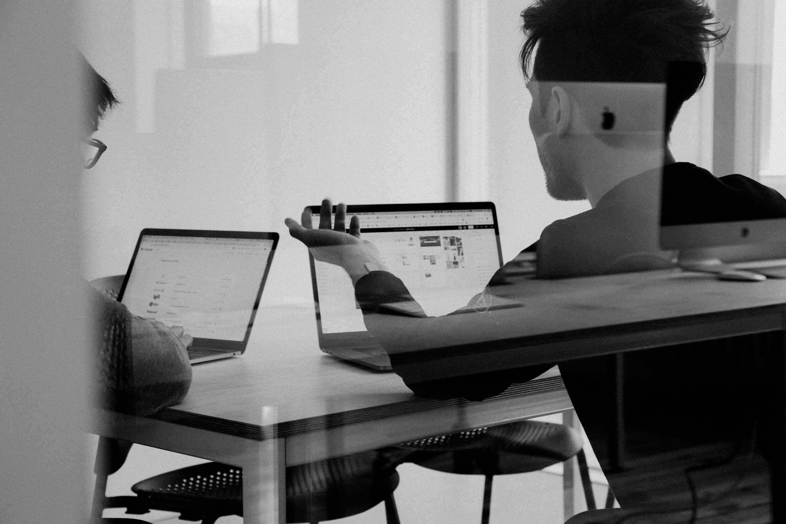 Two people working on laptops in a meeting room, viewed through glass.