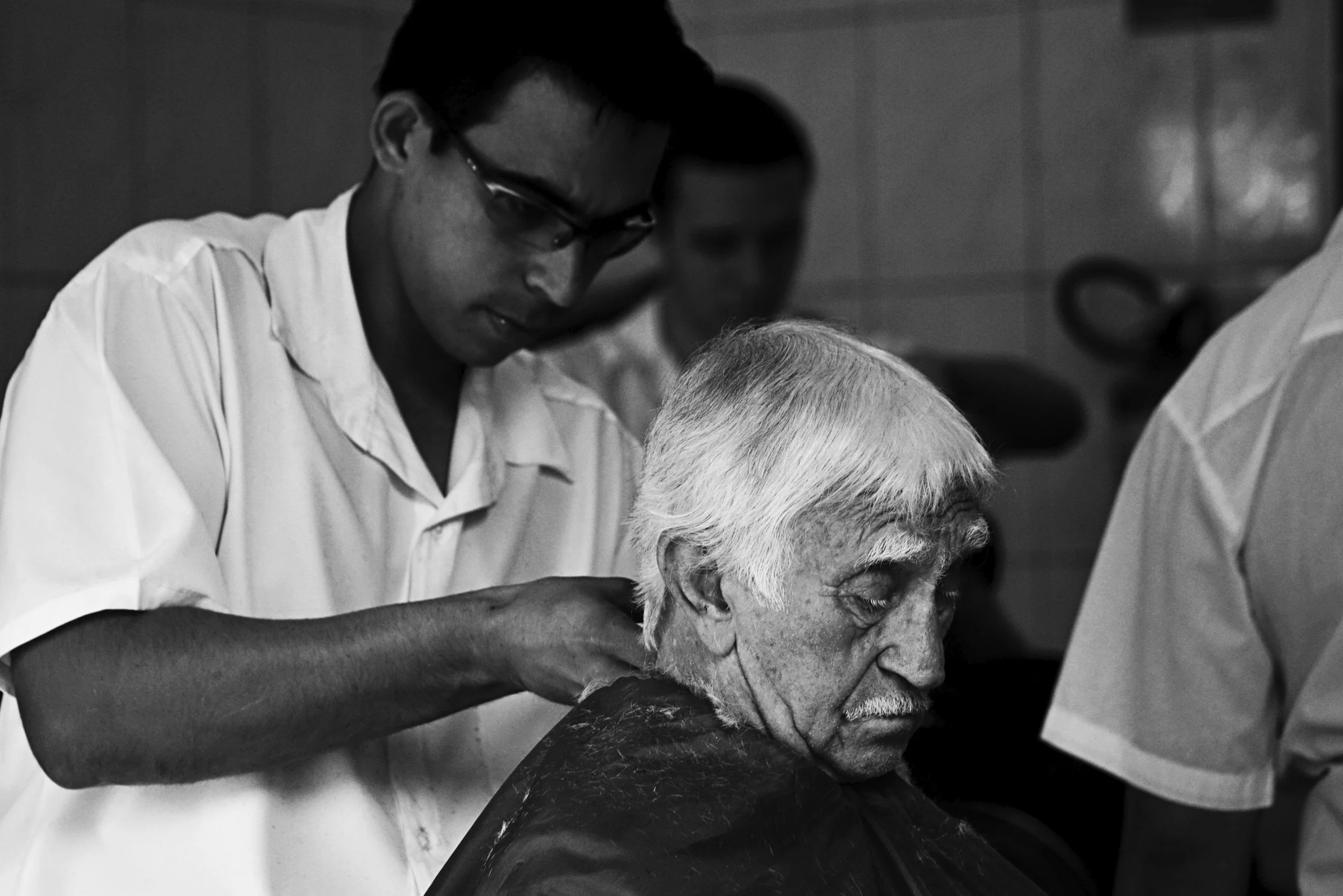 An elderly man with white hair and a mustache receives a haircut from a barber in a barbershop, with another person visible in the background.