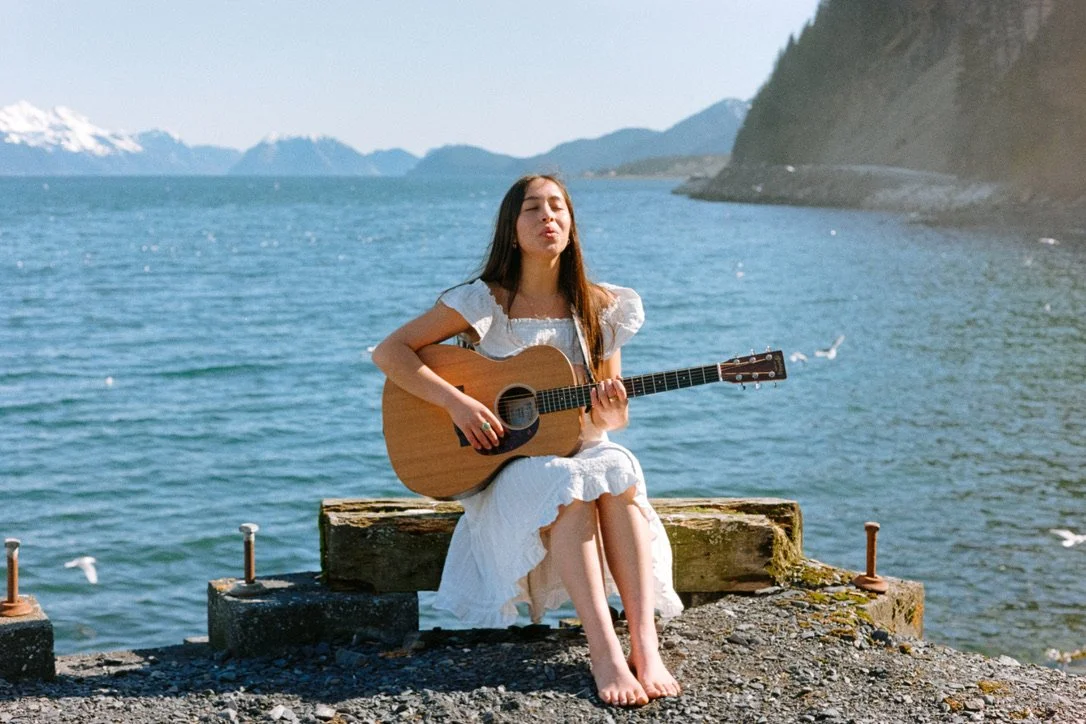 A woman in a white dress sitting on a concrete ledge by the water, playing an acoustic guitar, with mountains in the background.
