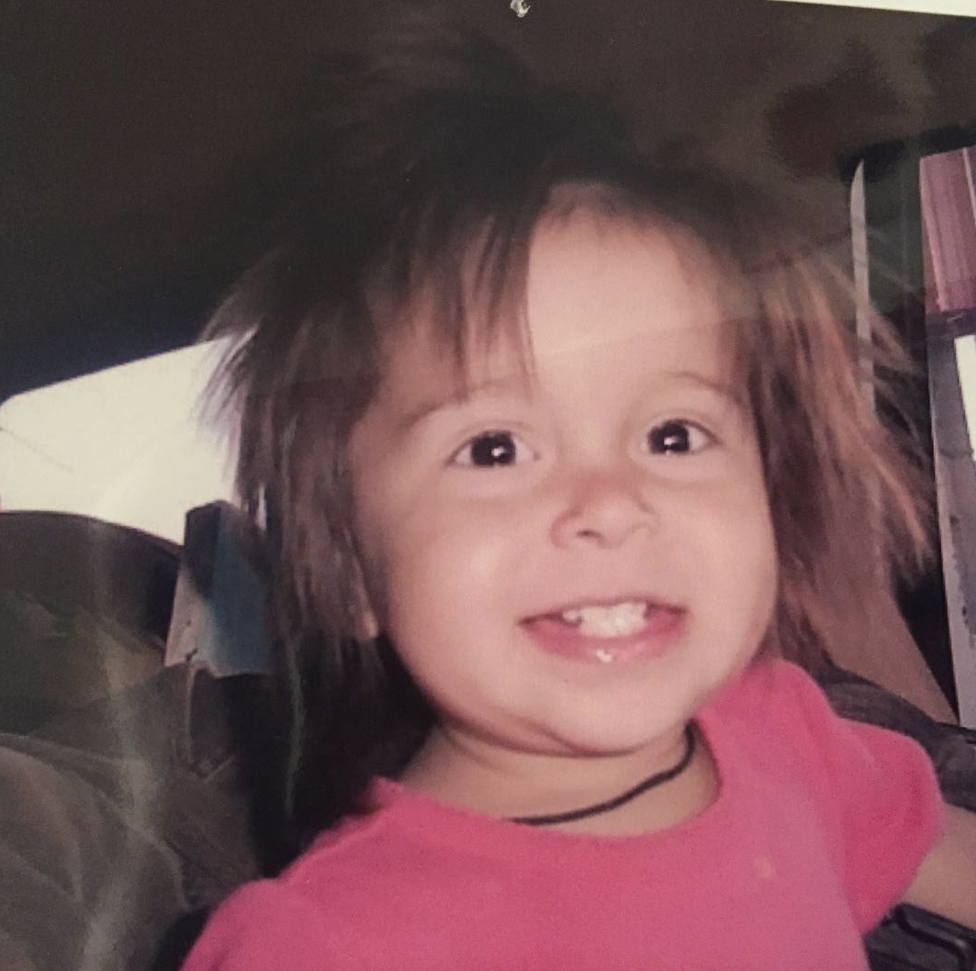 A young girl with brown hair, missing a front tooth, smiling, wearing a pink top and a black necklace, sitting in a car seat.