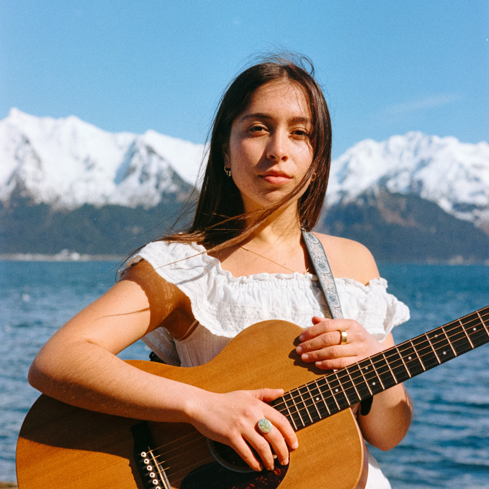 A woman with long brown hair holding an acoustic guitar in front of a mountain range with snow-capped peaks under a blue sky.